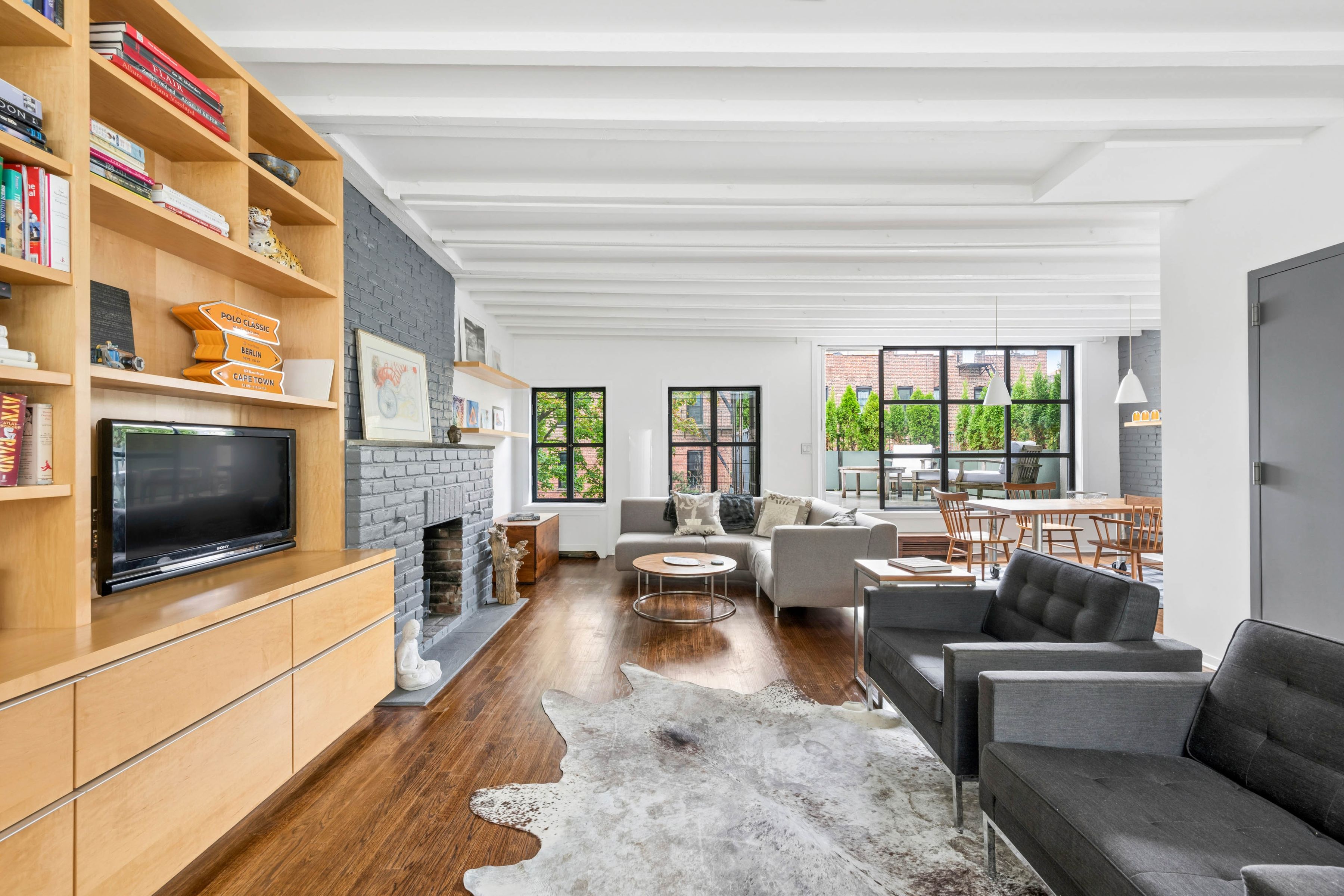 Open-plan living room with built-in shelving, gray brick fireplace, dark wood floors, and windows overlooking a terrace.