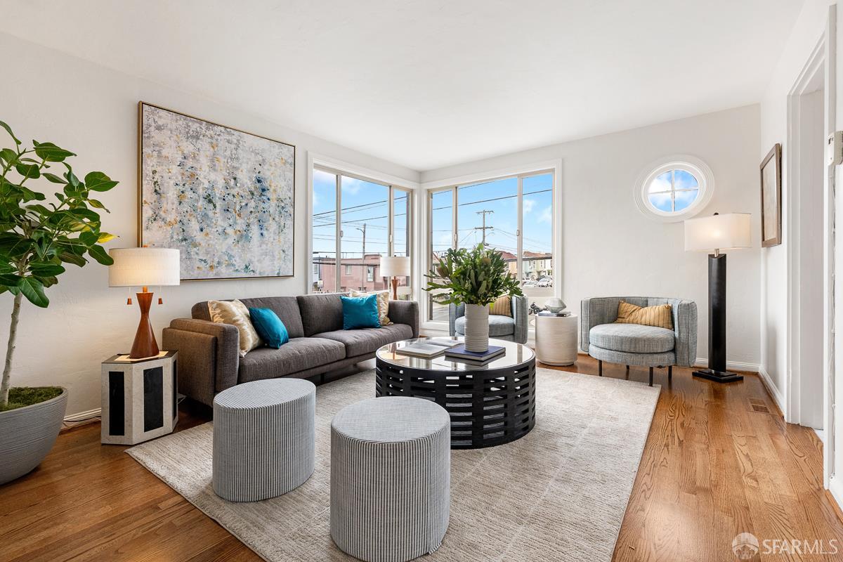 Staged living room with corner windows, gray seating, a round coffee table, and hardwood floors.