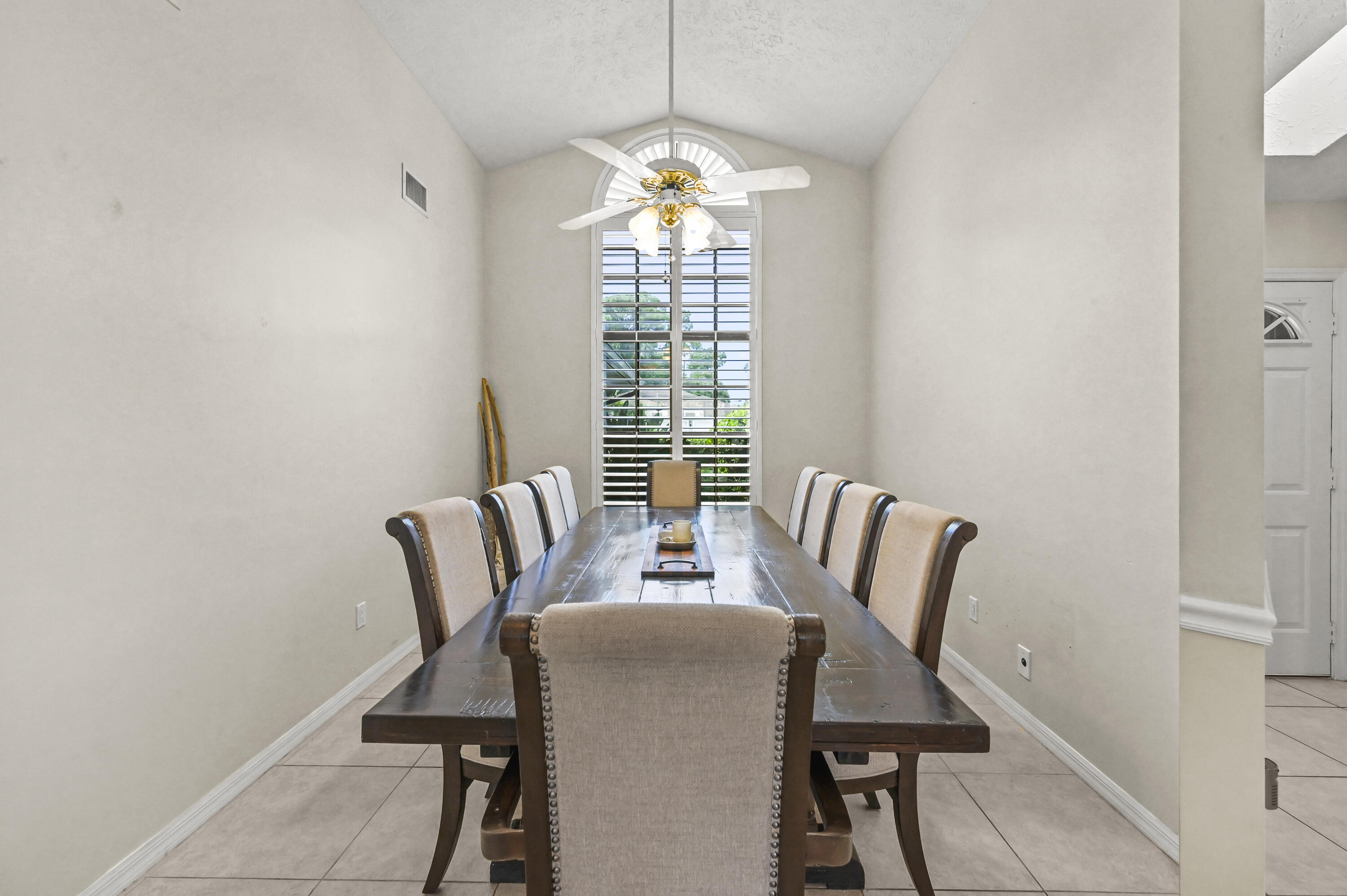 Furnished dining room with a long wood table beneath a vaulted ceiling and an arched shuttered window.