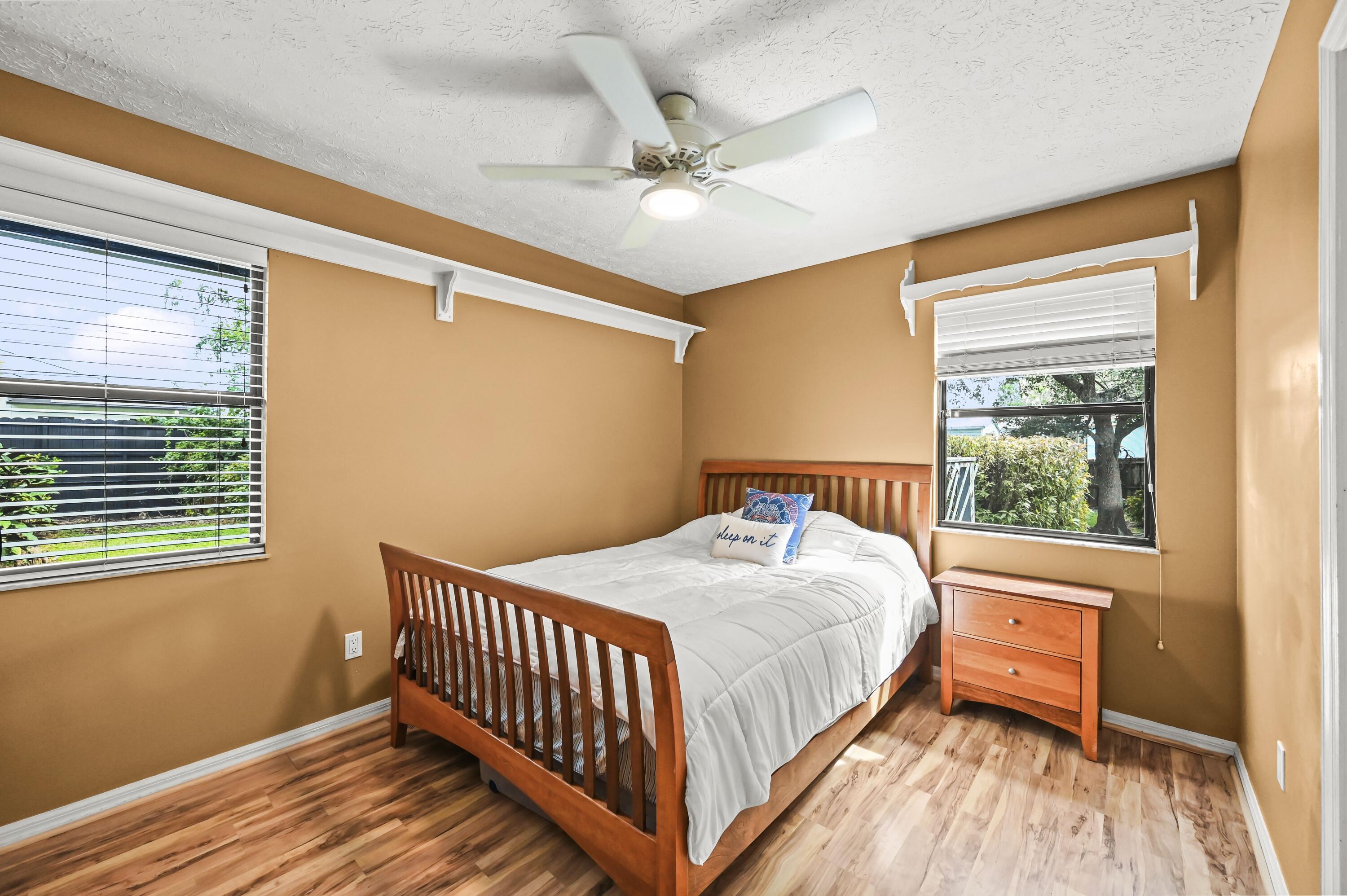 Furnished bedroom with a wooden bed, white bedding, tan walls, and windows with blinds.