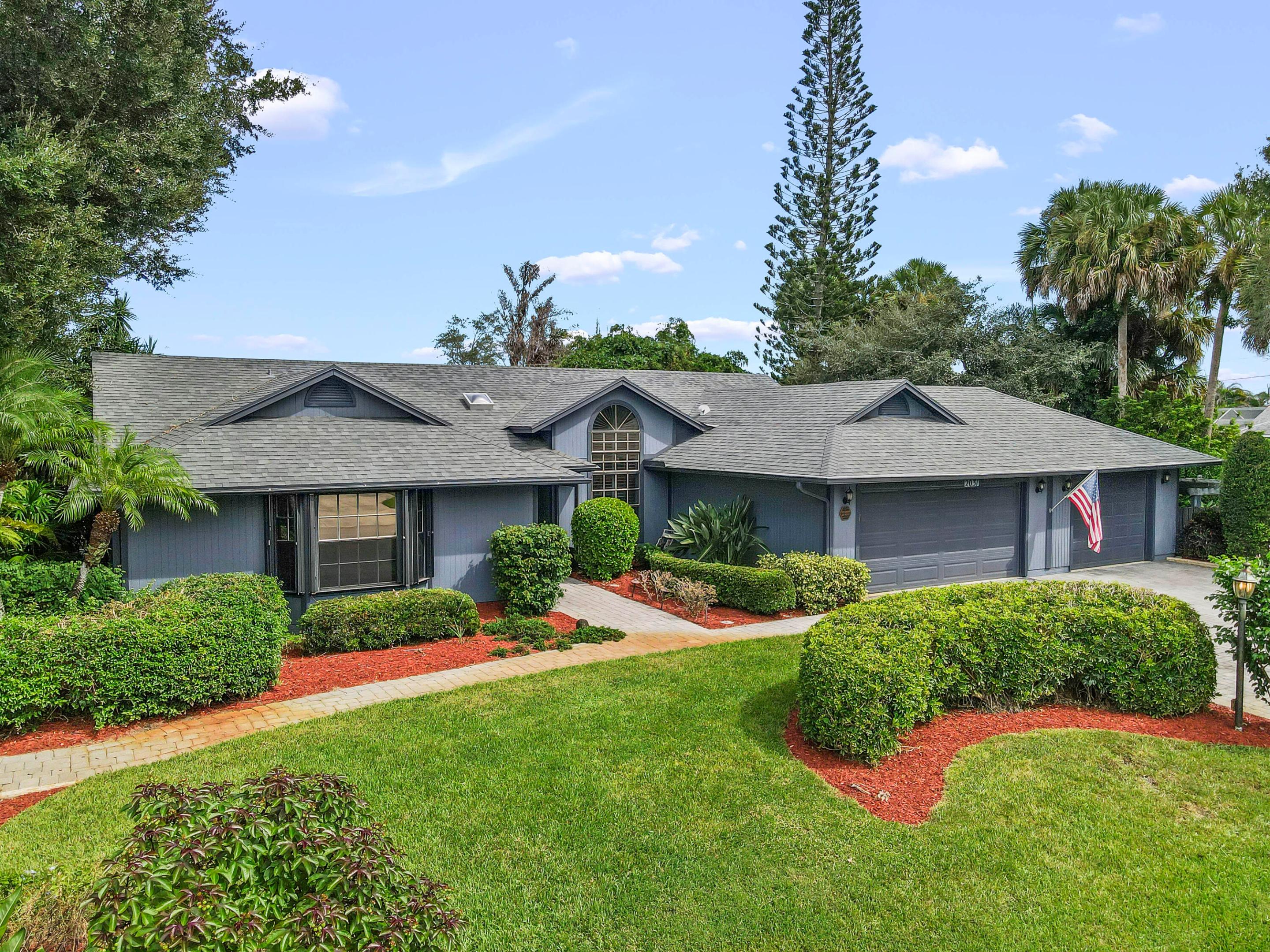 Blue-gray single-story home with an arched window, two-car garage, American flag, and landscaped front lawn.