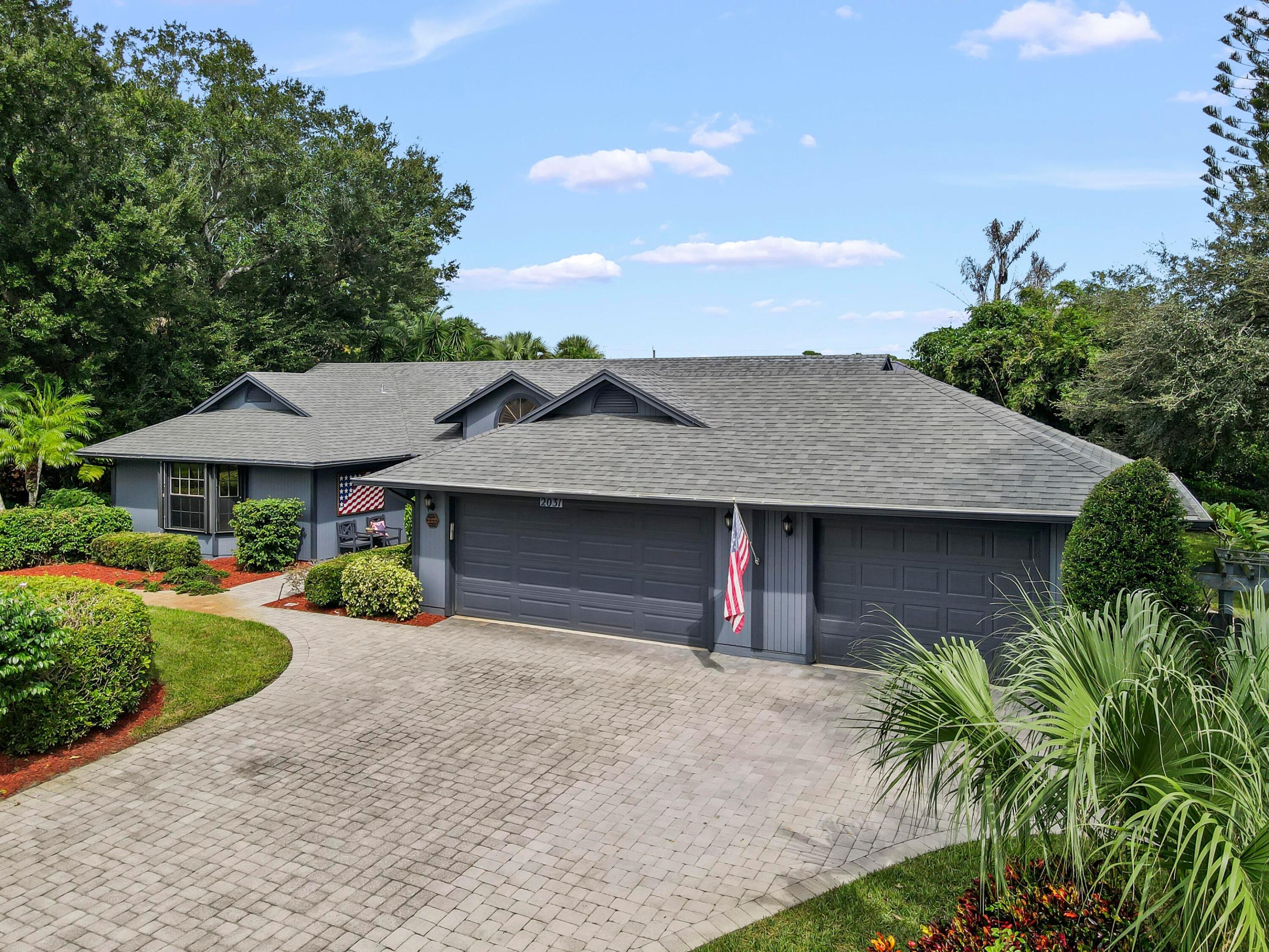 Gray single-story home with two garage doors, a wide paver driveway, and landscaped grounds.