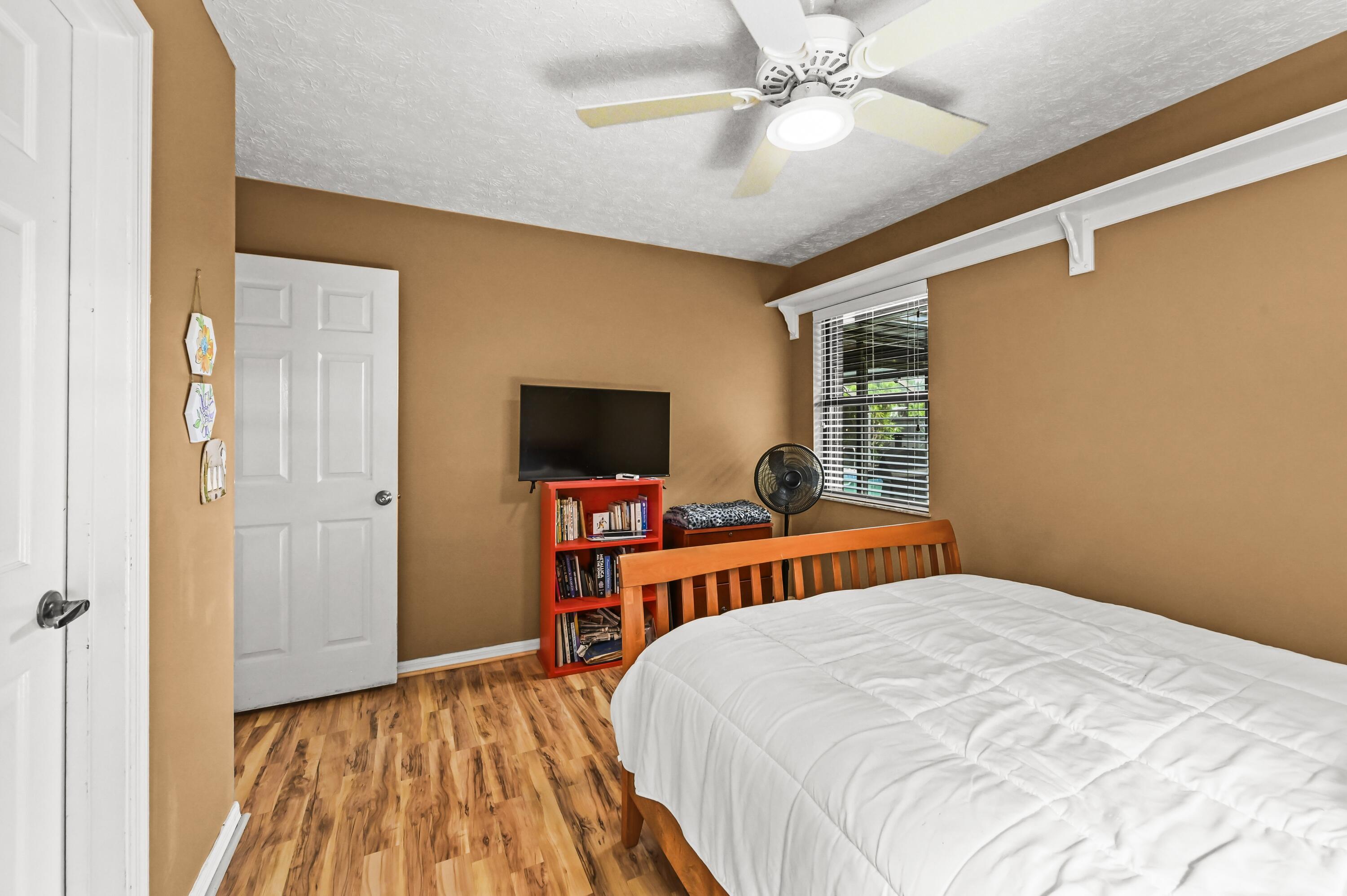 Bedroom with a white bed, tan walls, wood-look flooring, and a television on a red bookcase.