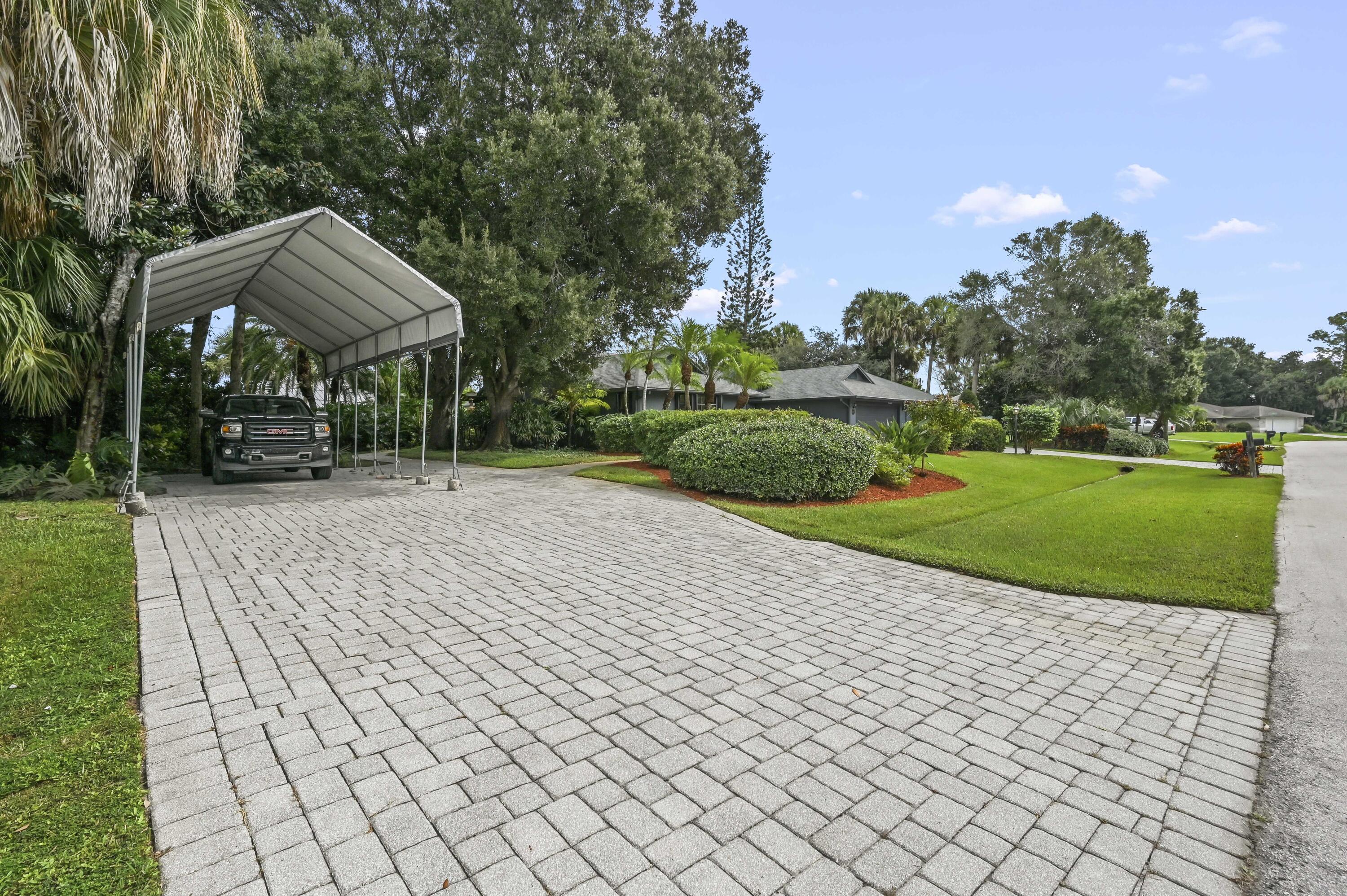 Home behind a wide paver driveway, with a metal carport and tropical landscaping.