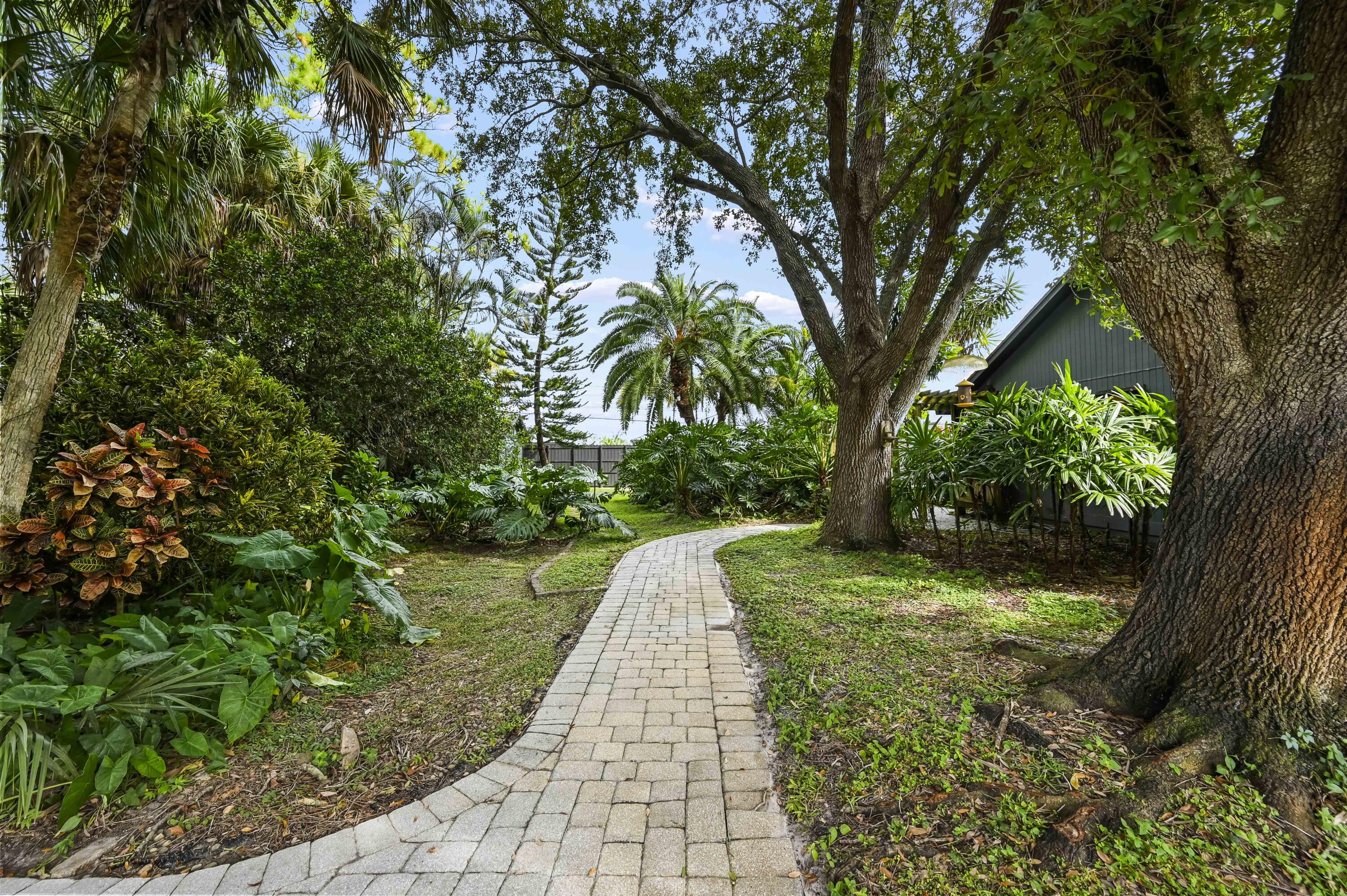 Curving paver walkway through a tropical garden beside a dark-sided house and mature trees.