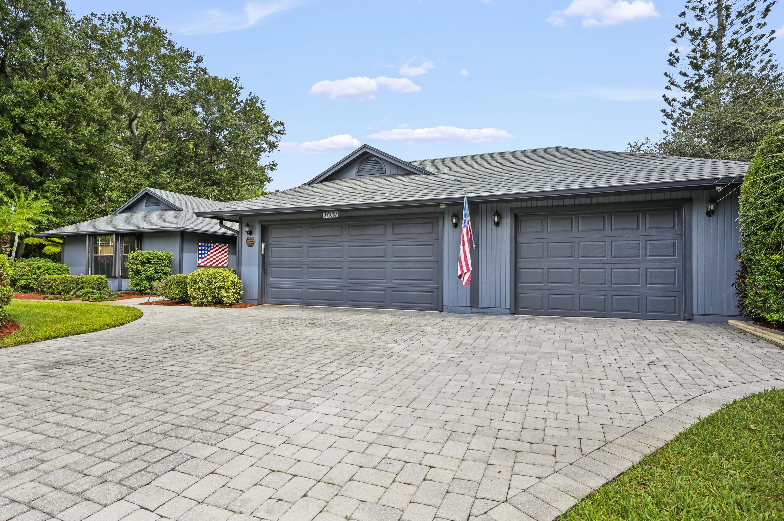 Blue-gray home with two garage doors, a wide paver driveway, and landscaped front yard.