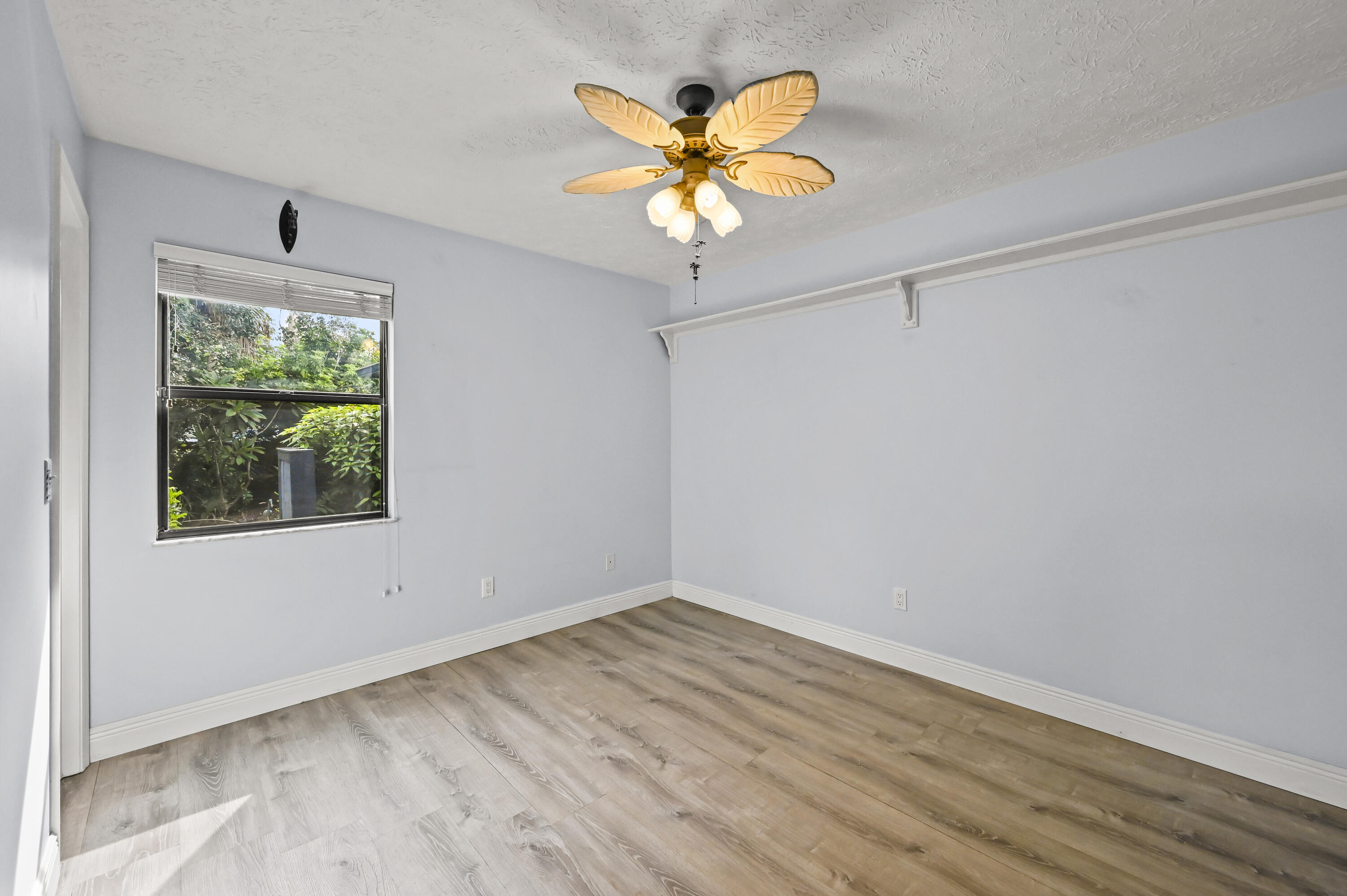 Empty bedroom with wood-look flooring, a leafy window view, and a lit leaf-blade ceiling fan.