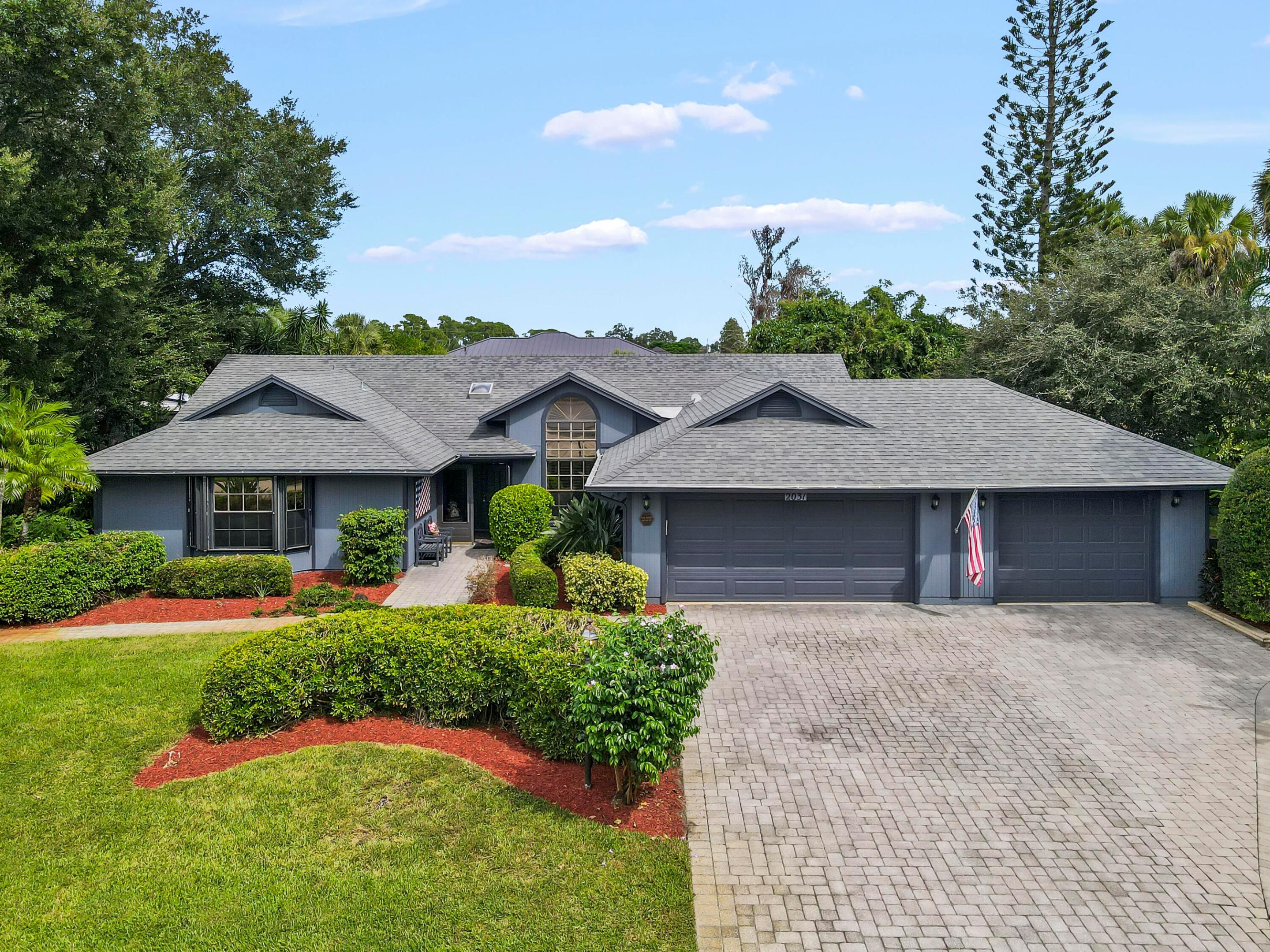 Blue-gray home with a two-car garage, wide paver driveway, landscaped lawn, and American flag.