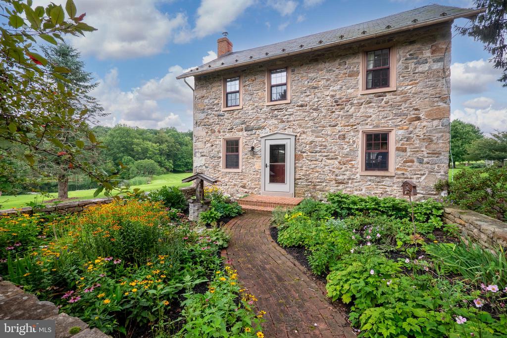 Stone house with a curved brick walkway through colorful flower gardens.