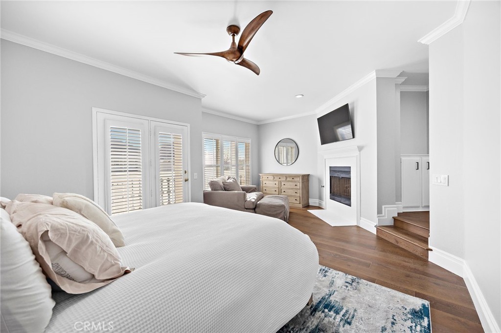 Bedroom with a large white bed, plantation shutters, and a fireplace with a television above it.