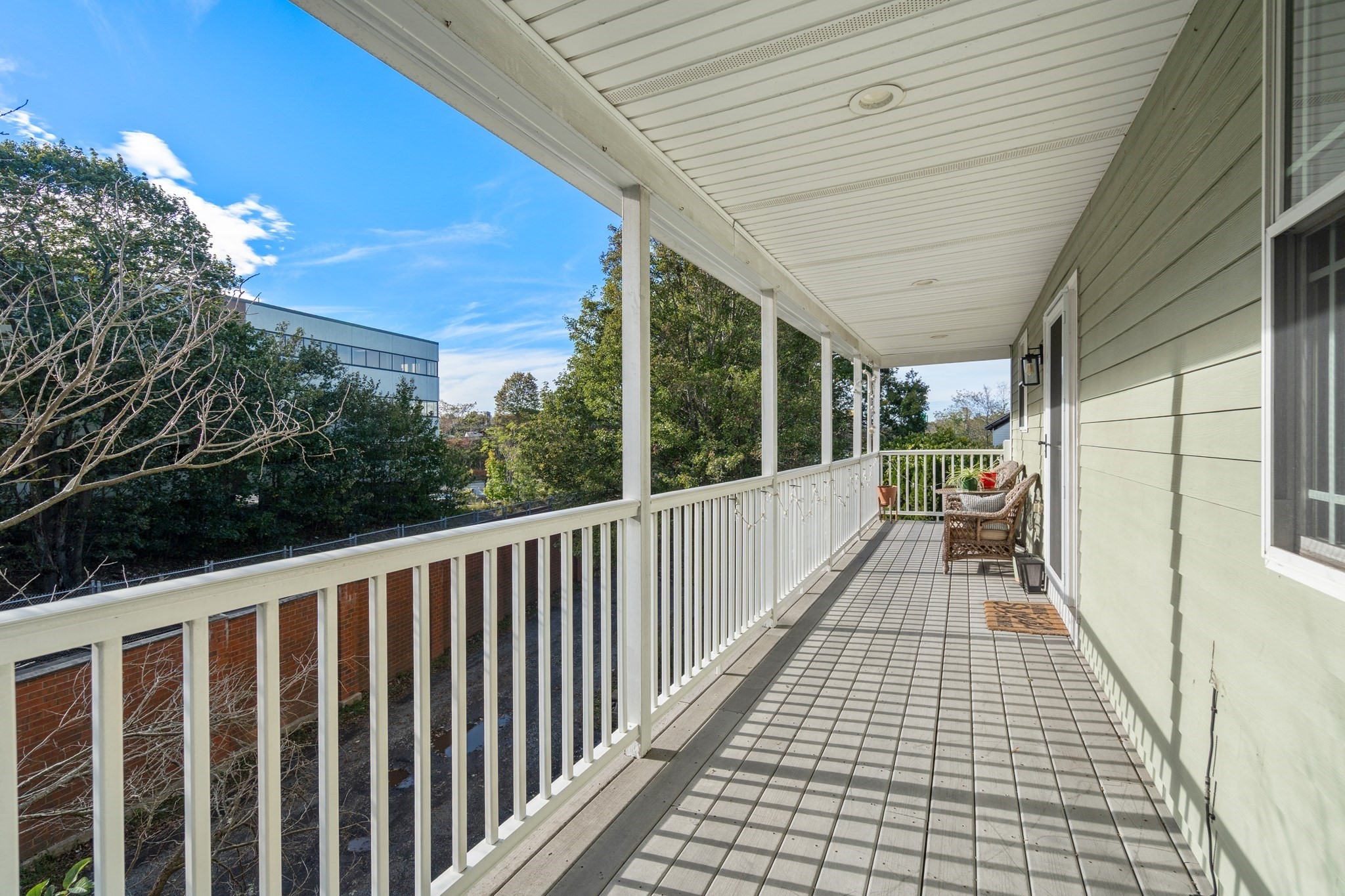 Covered porch with white railings, gray plank flooring, and wicker seating overlooking trees.