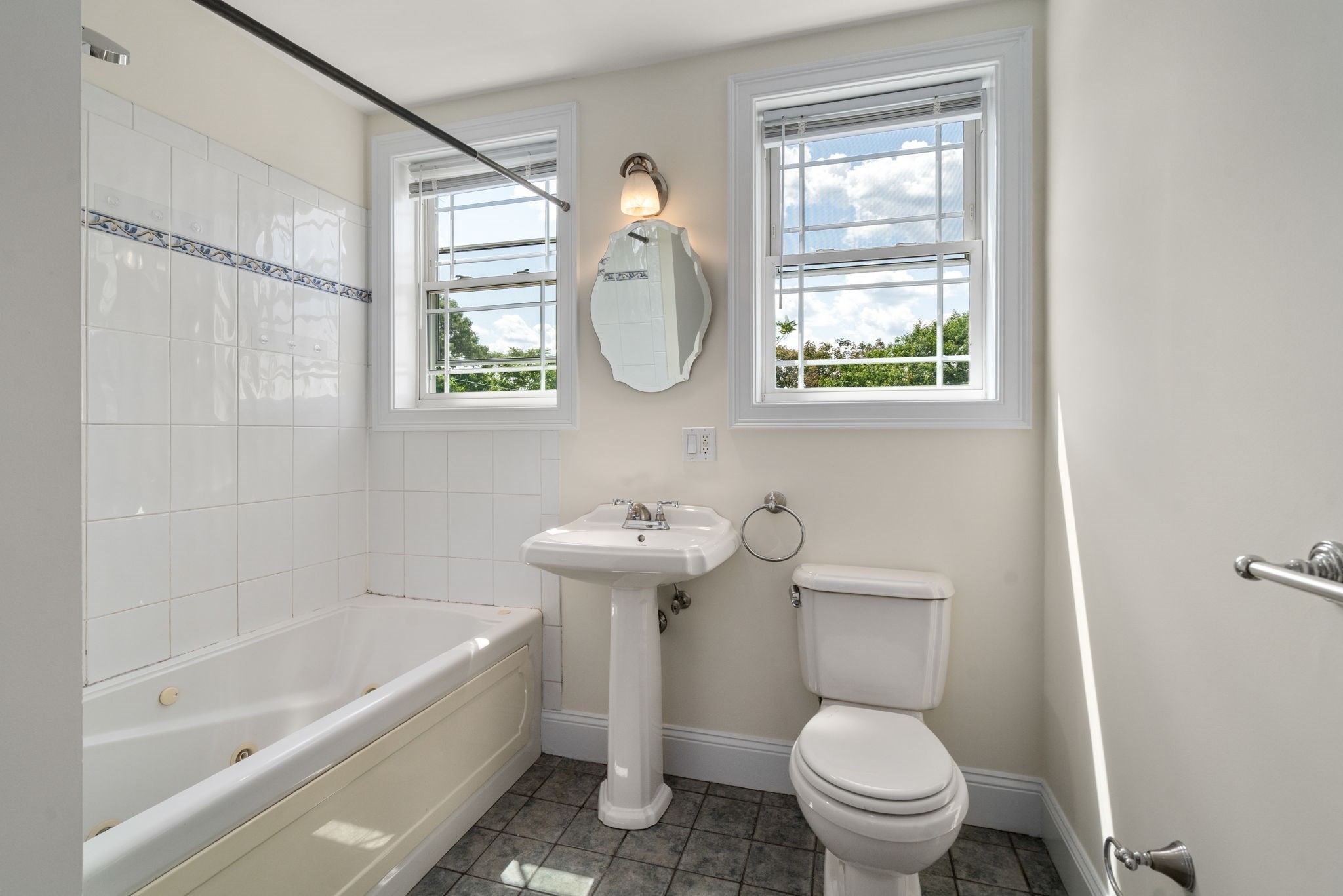 Bathroom with a tiled bathtub, pedestal sink, toilet, and two sunny windows.