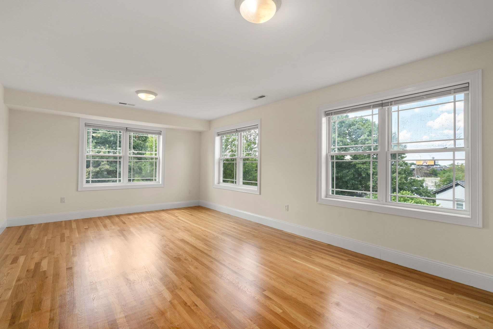 Empty room with hardwood floors and three large windows overlooking trees.