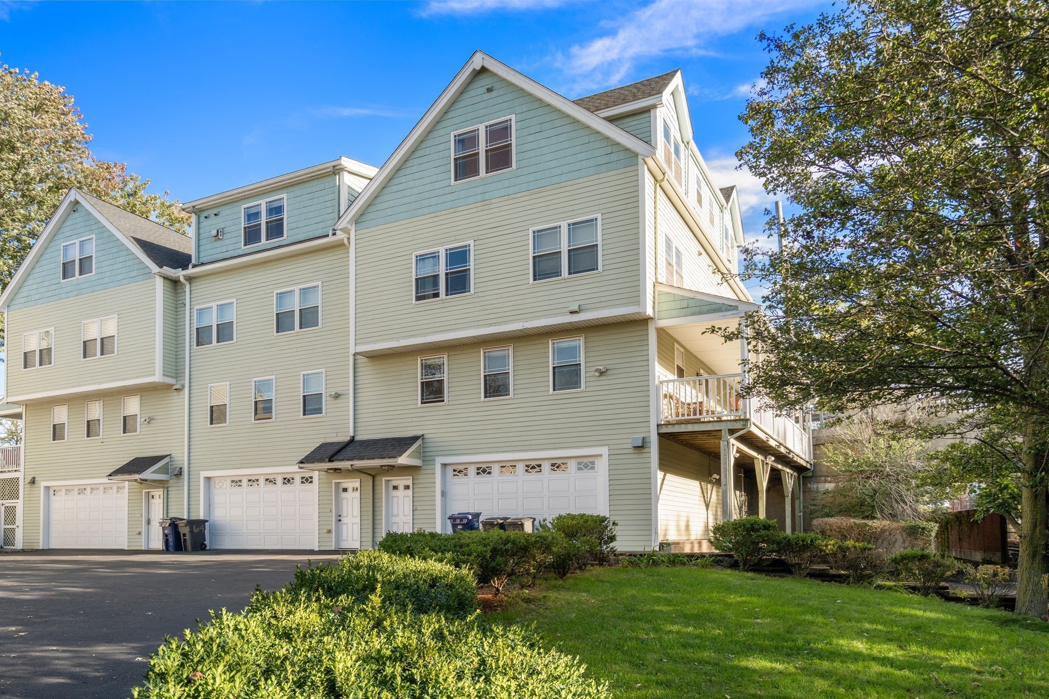Multi-story sage-green residence with white garage doors, a raised balcony, and a landscaped lawn.