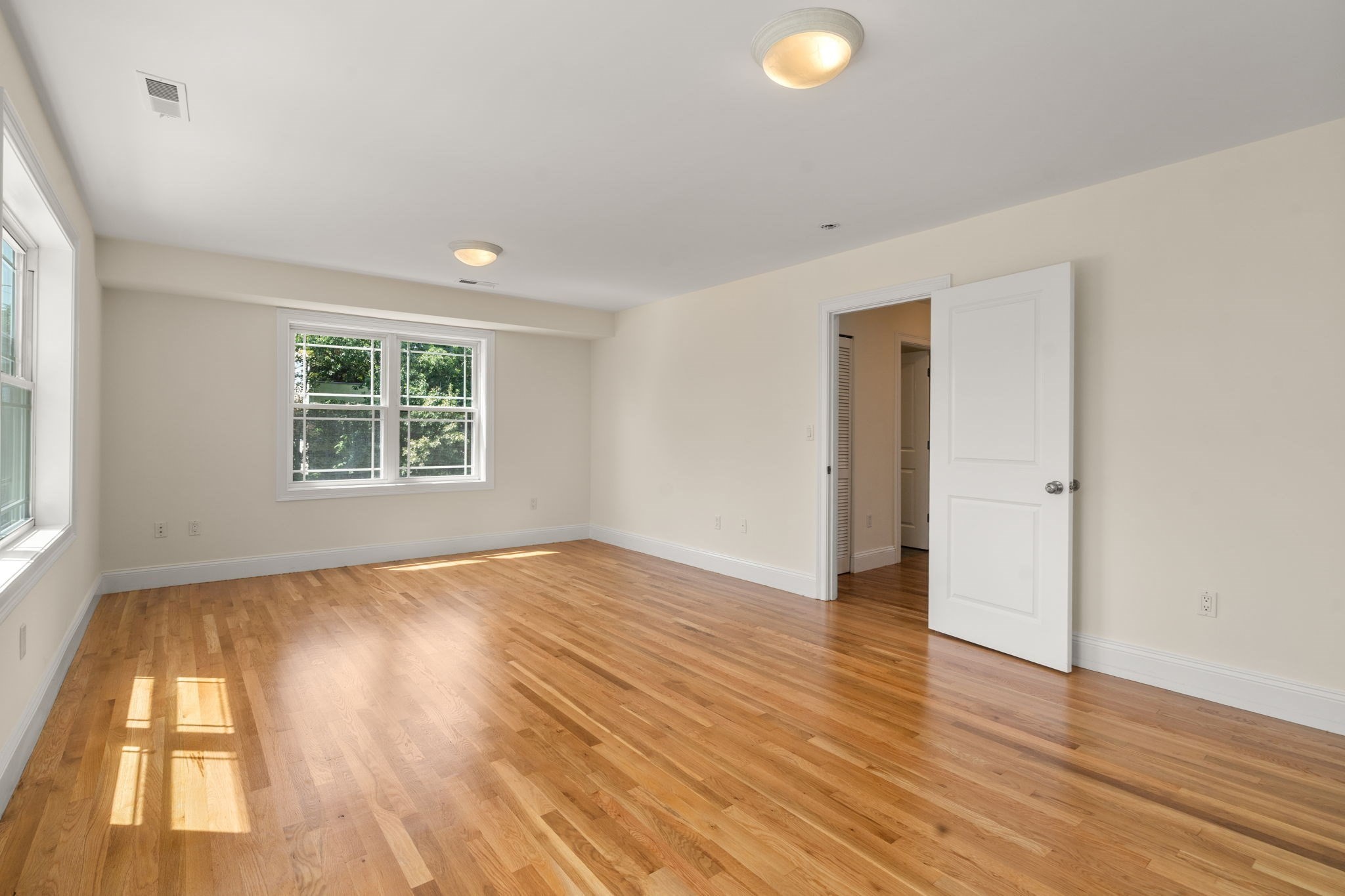 Empty bedroom with polished hardwood floors, two windows, and sunlight across the floor.