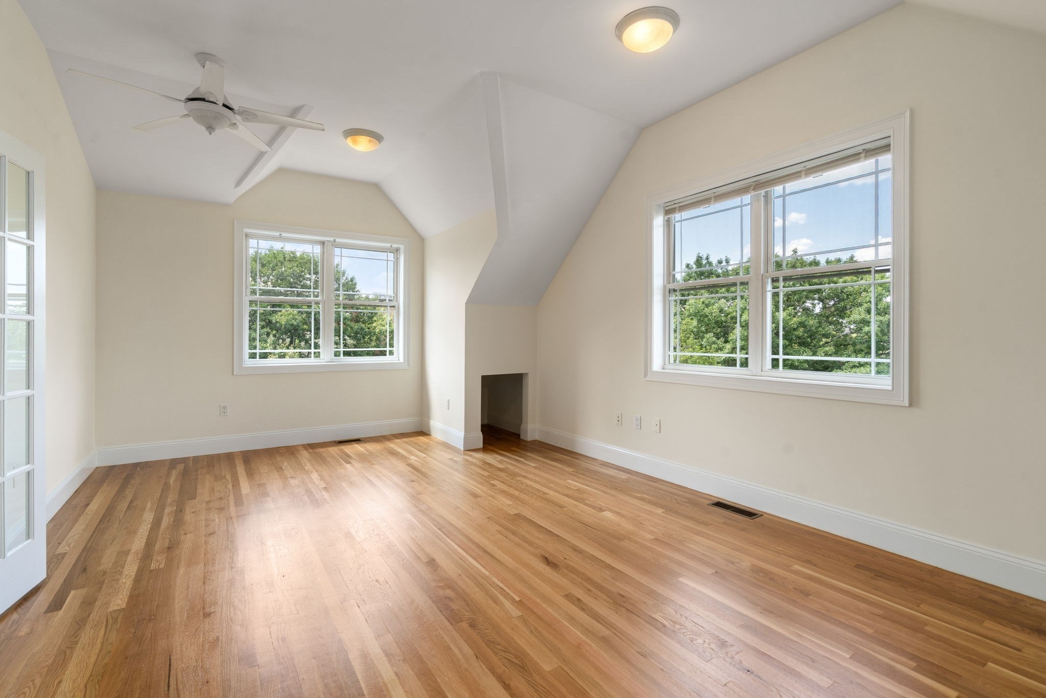 Empty room with vaulted ceiling, hardwood floors, ceiling fan, and large windows overlooking trees.