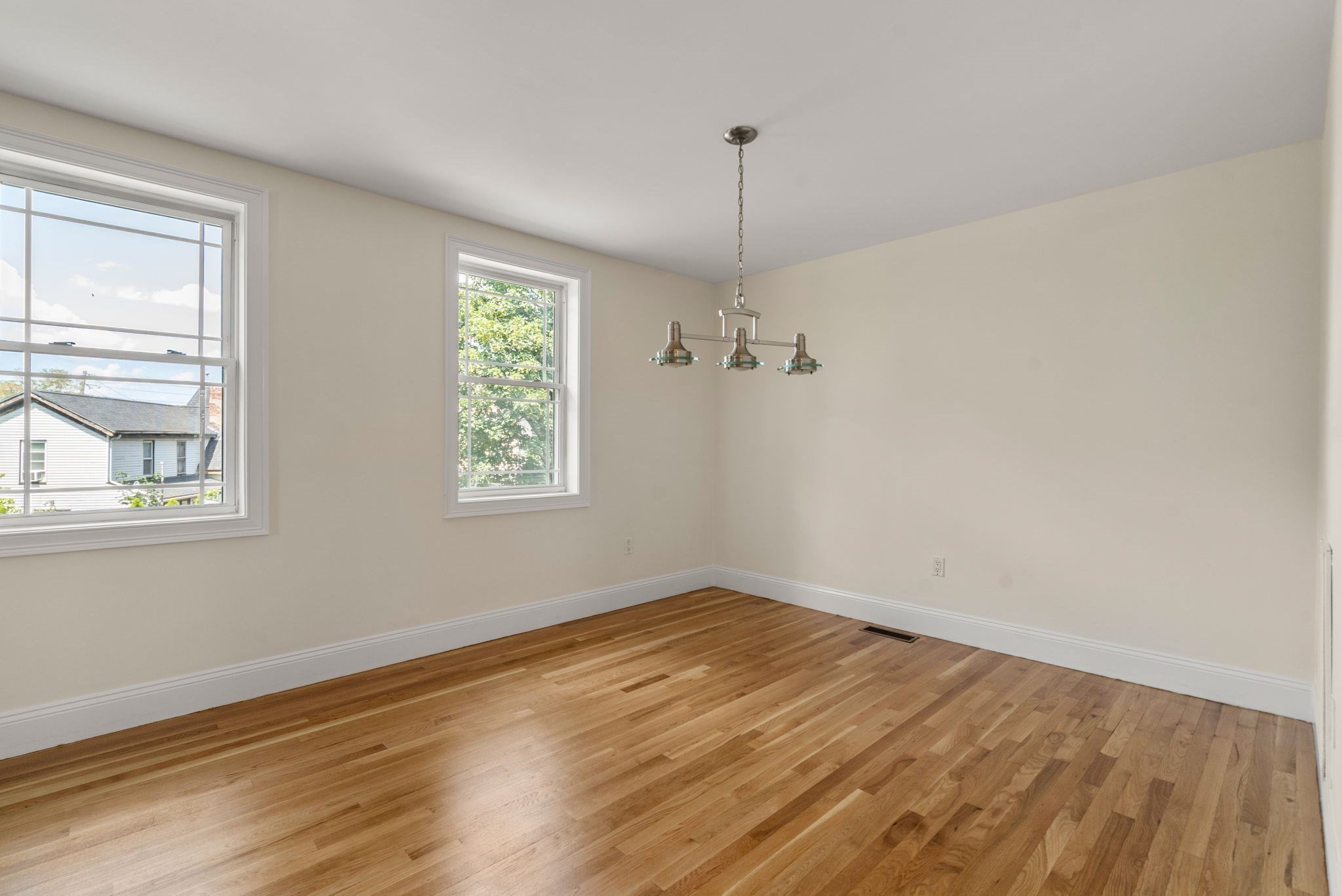 Empty dining room with polished hardwood floors, two windows, and a hanging three-light fixture.