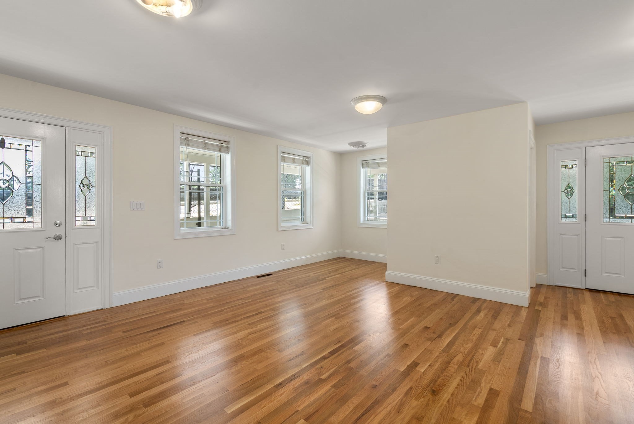 Empty living room with hardwood floors, multiple windows, and decorative stained-glass entry doors.