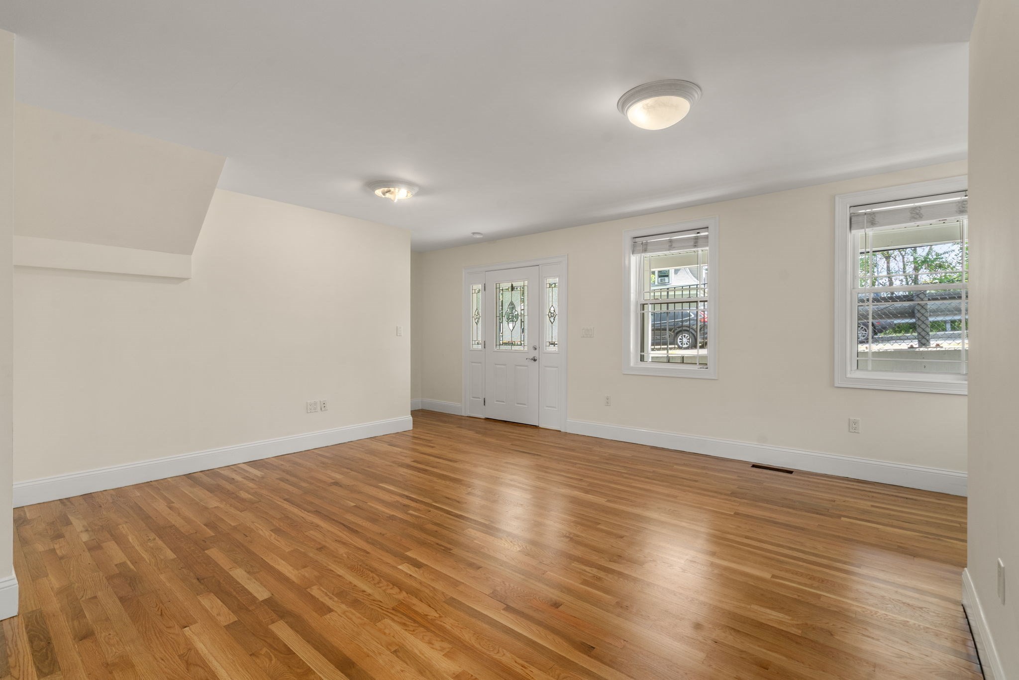 Empty living room with polished hardwood floors, two windows, and a decorative glass front door.