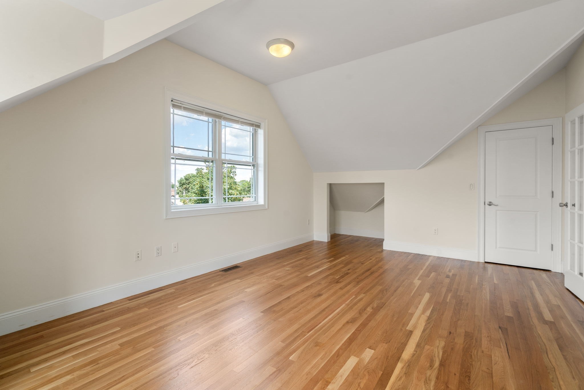 Empty room with sloped ceilings, polished hardwood floors, and a large window.
