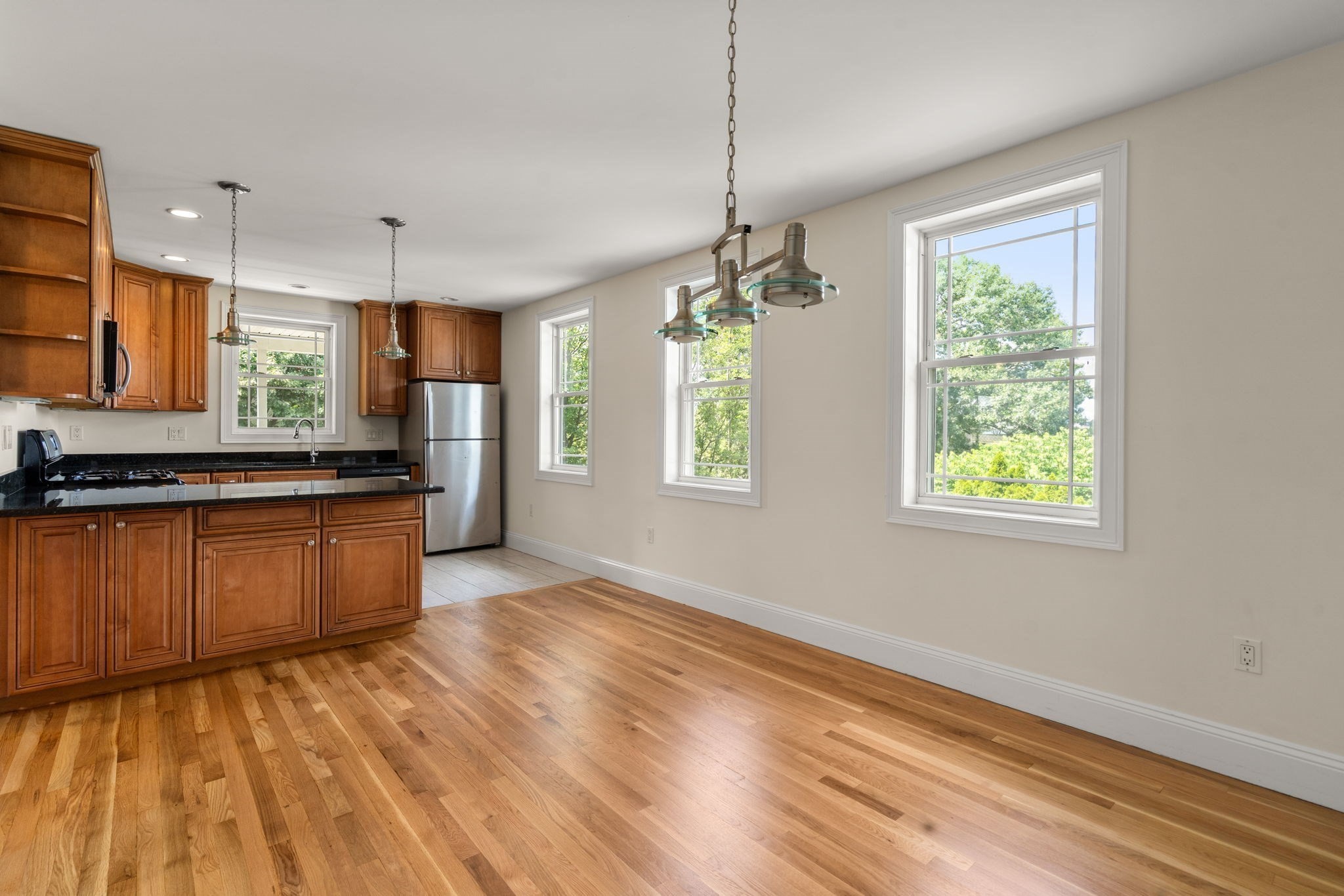 Open kitchen and dining area with wood cabinets, hardwood floors, and large windows facing trees.