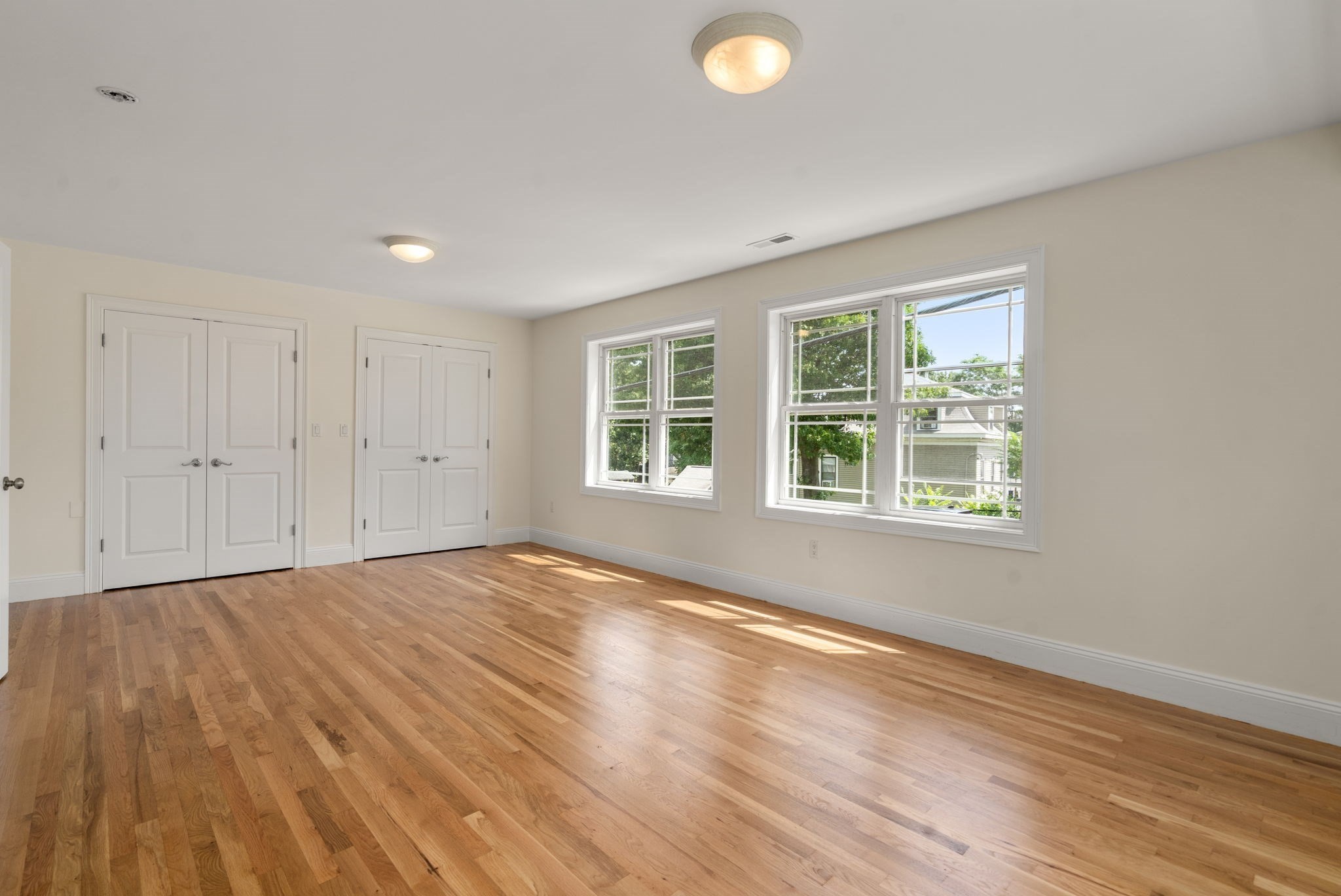Empty bedroom with hardwood floors, two sets of closet doors, and large sunlit windows.