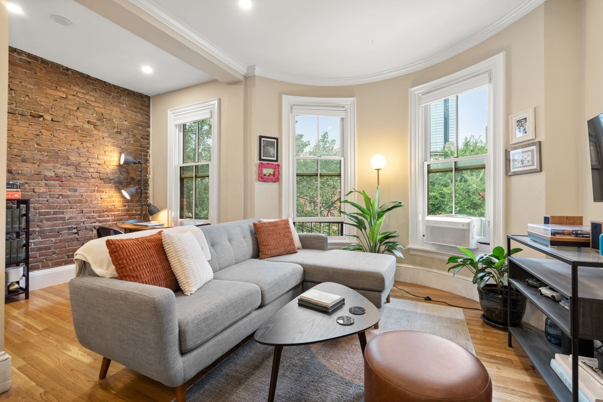 Living room with a gray sectional, exposed brick wall, and three tall windows.