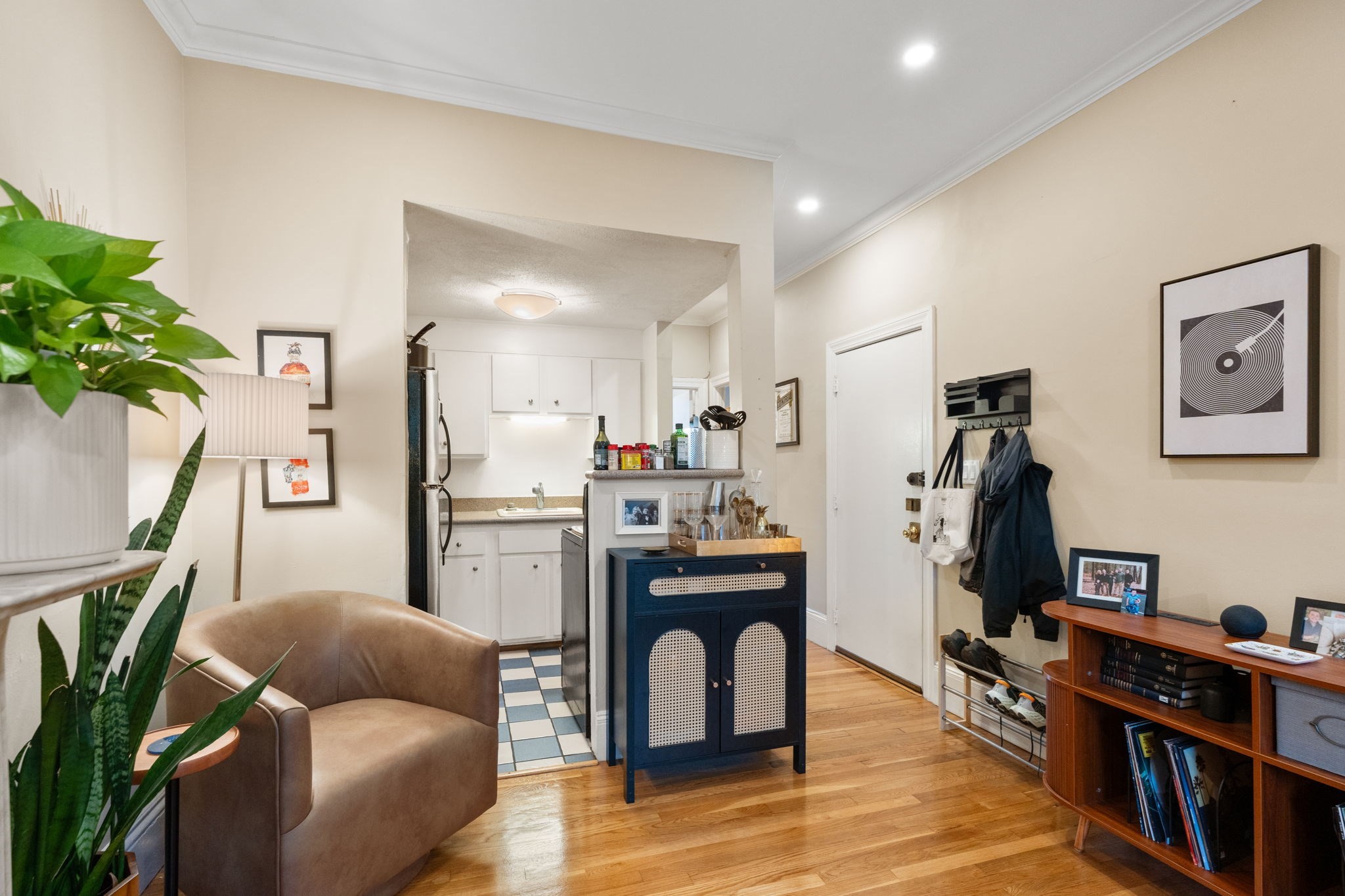 Furnished living room opening to a compact kitchen, with hardwood floors, leafy plants, and a dark blue cabinet.