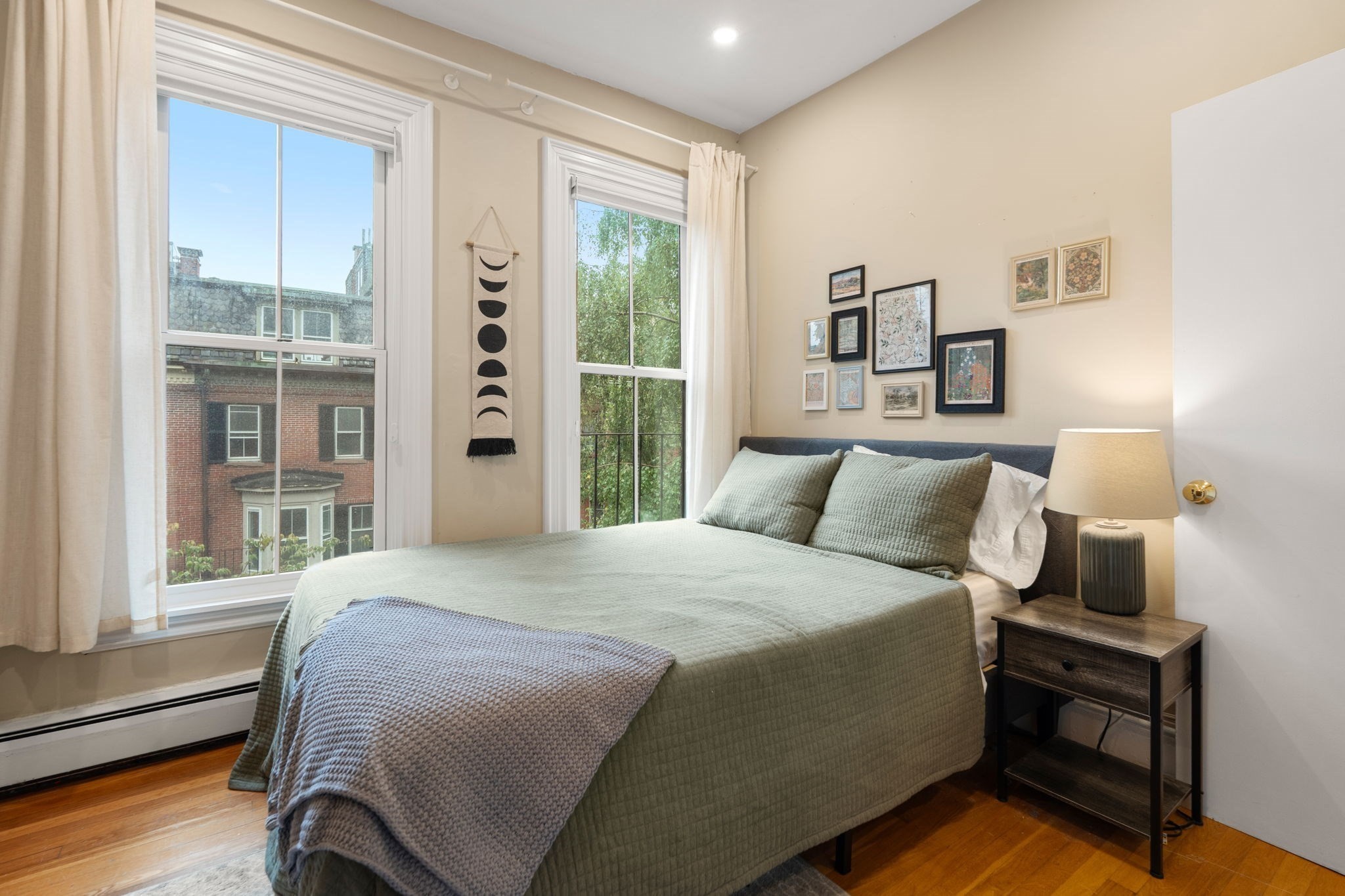 Bedroom with a sage-green bed, framed wall art, and two tall windows overlooking nearby buildings and trees.