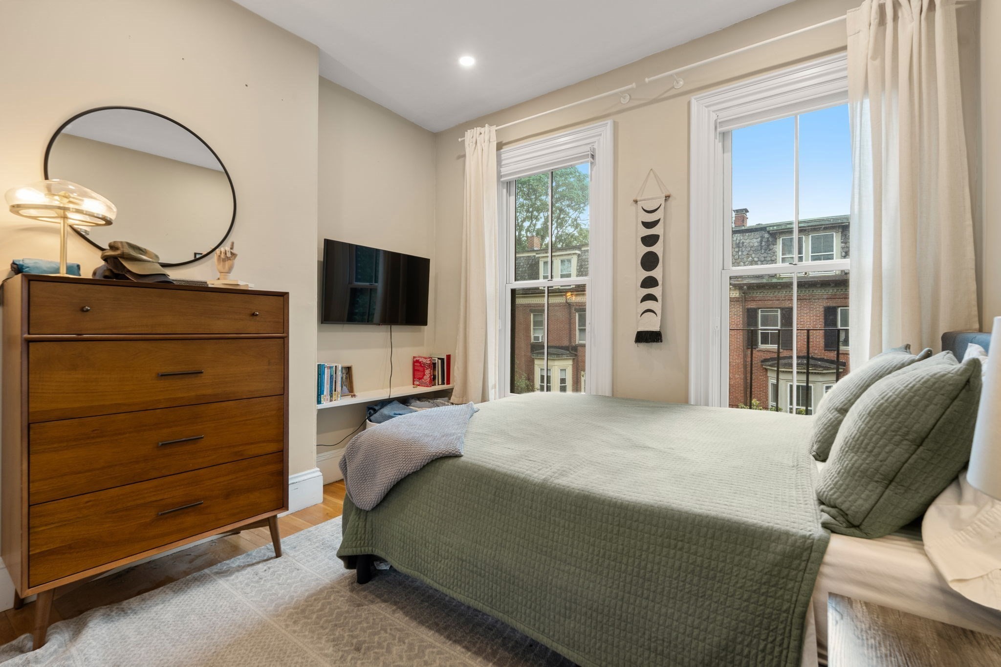 Furnished bedroom with a green quilted bed, wooden dresser, and two tall windows overlooking brick buildings.