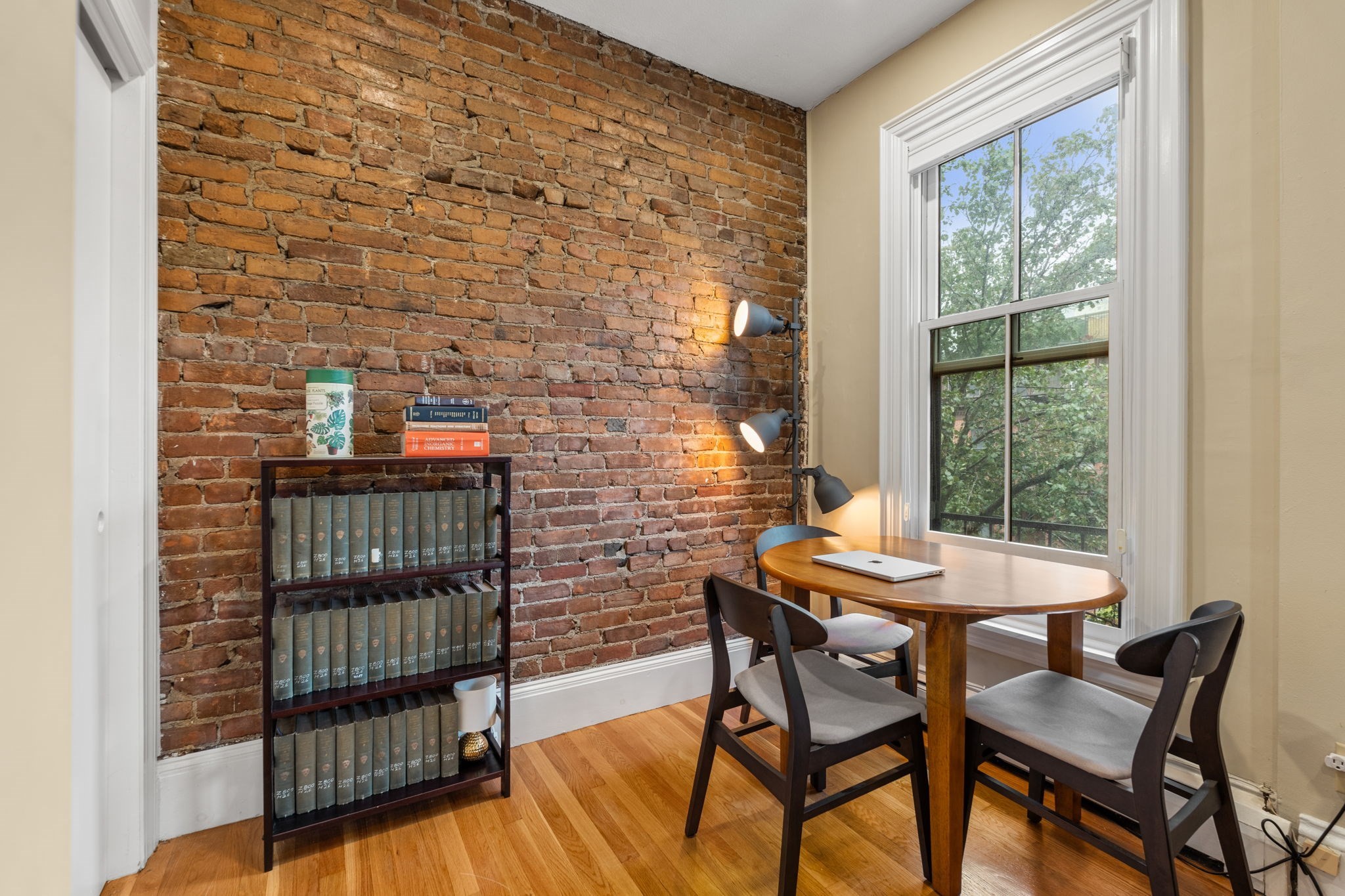 Furnished study with an exposed brick wall, bookcase, and round table beside a large window.