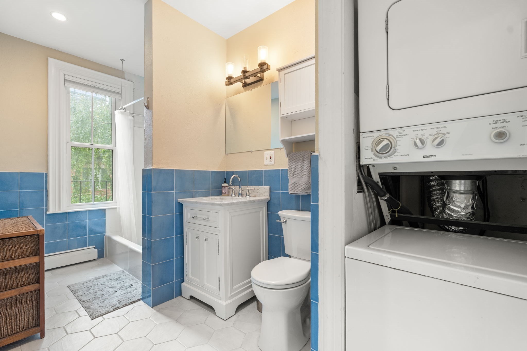 Bathroom with blue tile, a white vanity, window-side tub, and stacked washer and dryer.