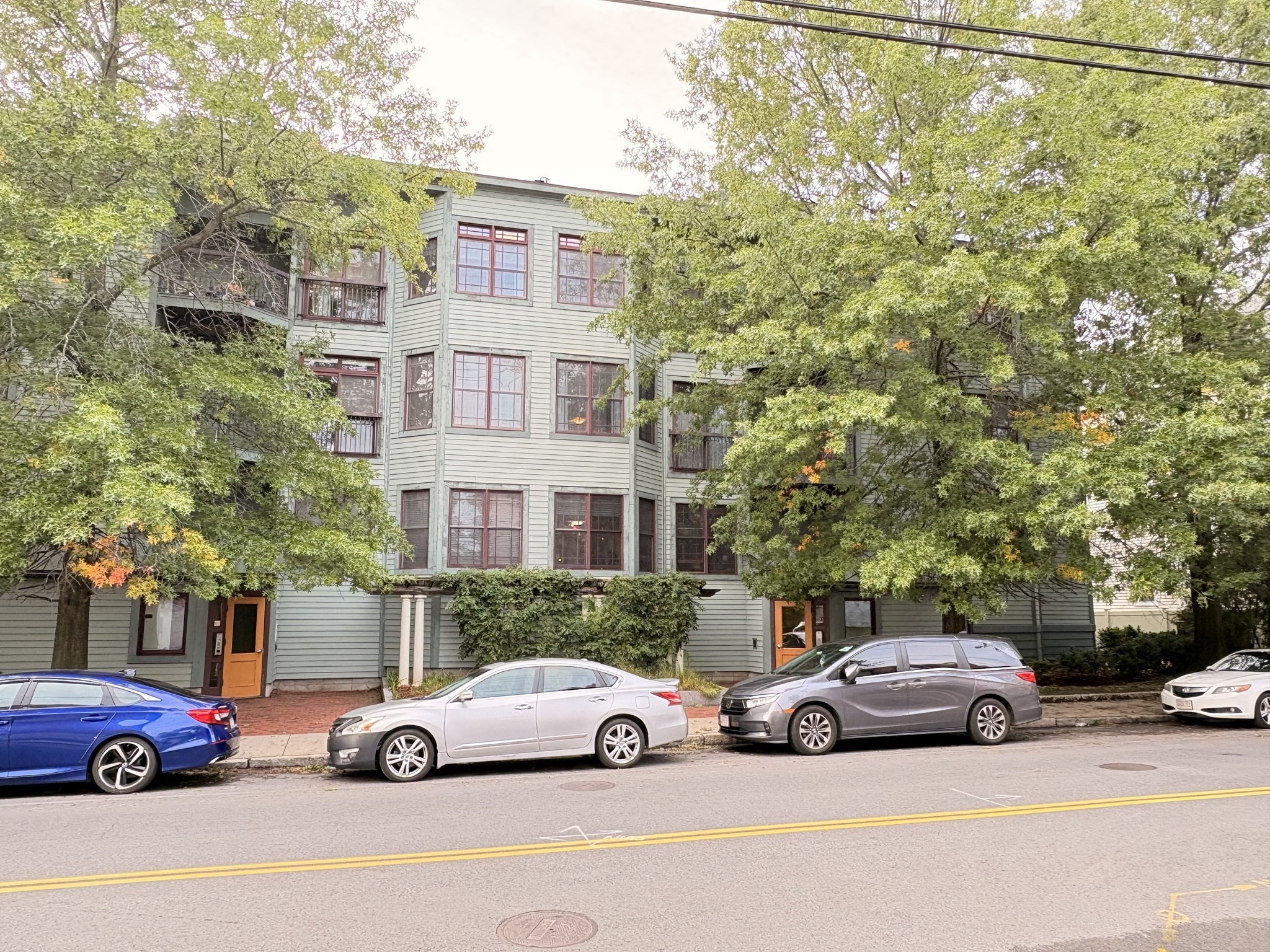 Multi-story residential building with red-trimmed windows and balconies behind leafy trees.