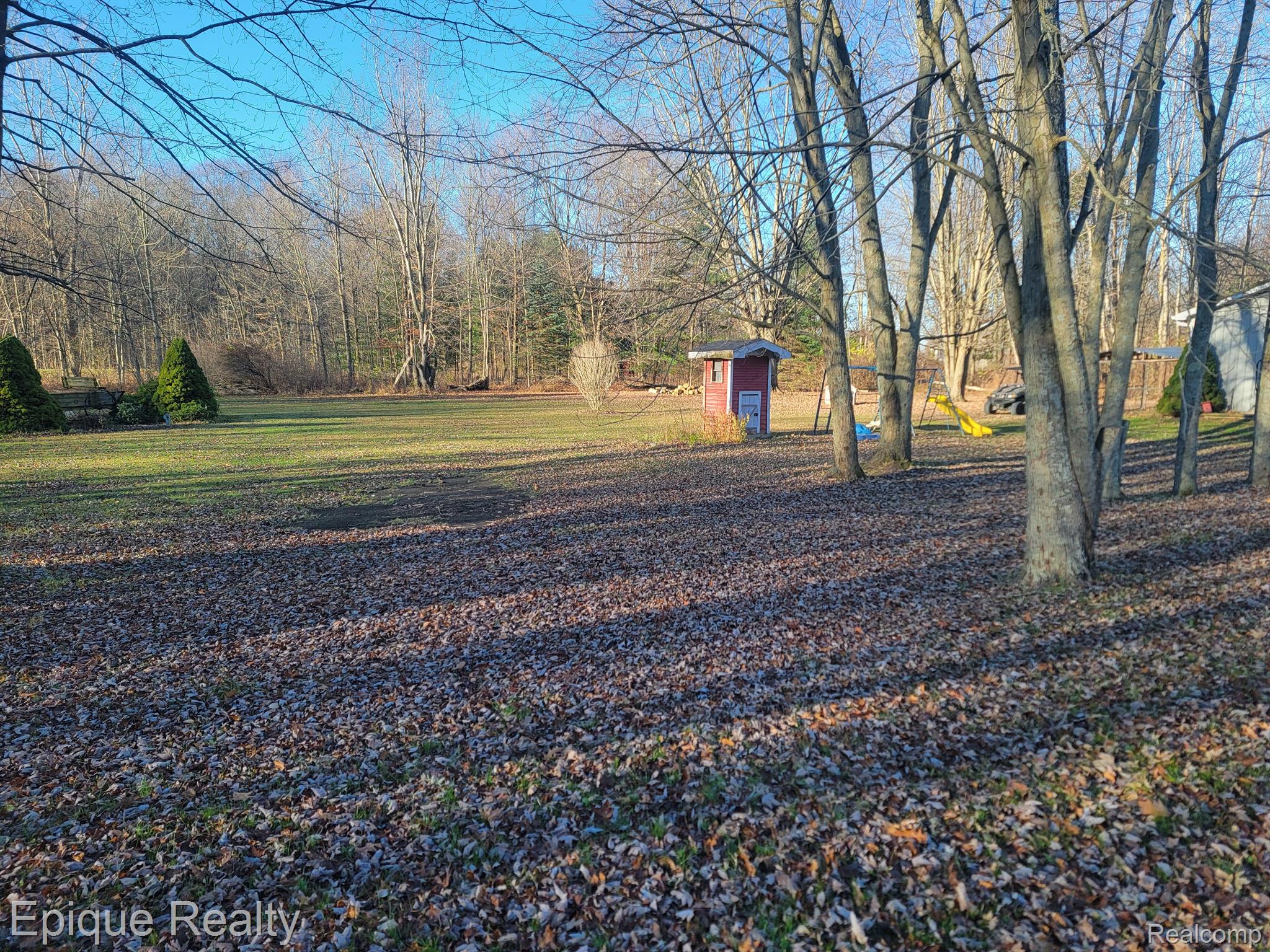 Backyard with mature trees, a red playhouse, yellow slide, and leaf-covered lawn.