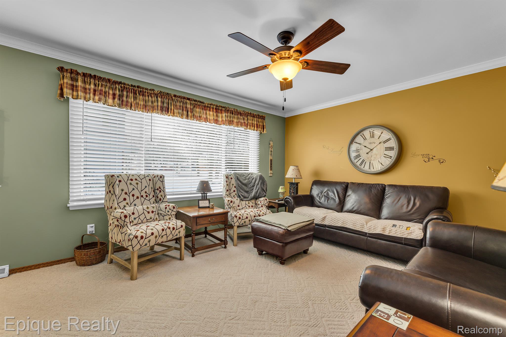Living room with patterned armchairs, dark leather seating, a large wall clock, and a lit ceiling fan.