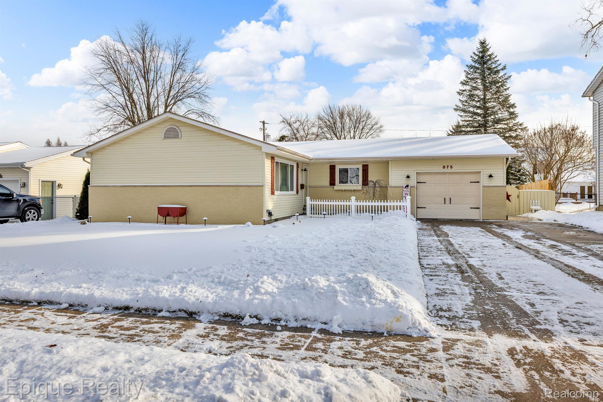 Cream-colored single-story home with attached garage, white picket fence, and snow-covered driveway.