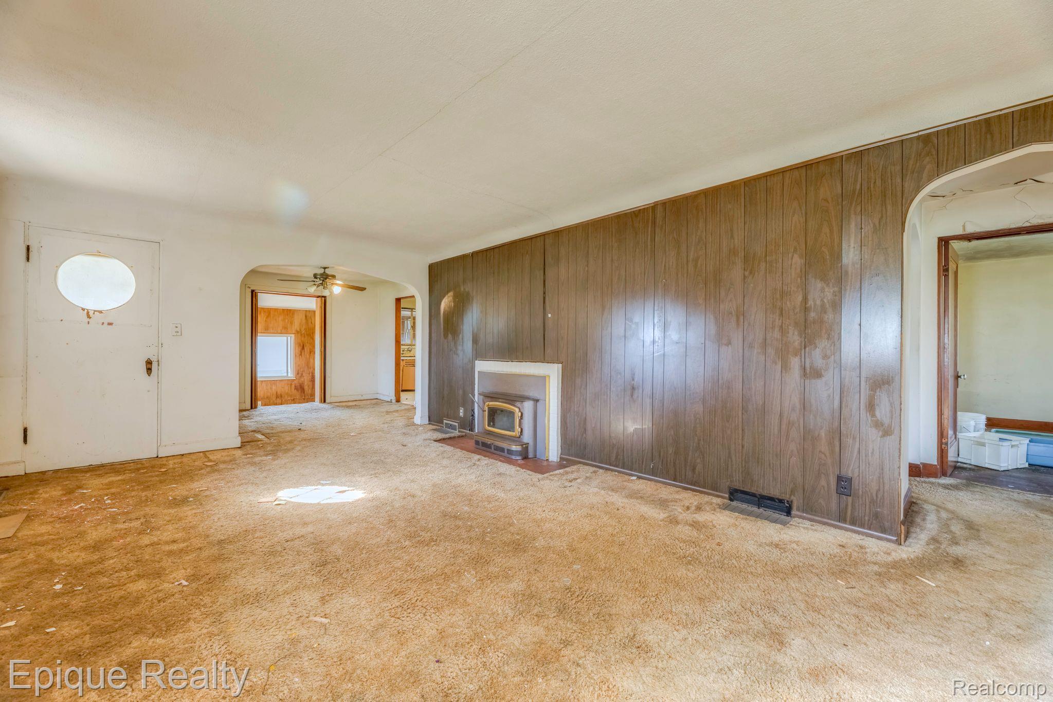 Vacant living room with wood-paneled walls, worn carpet, and a small fireplace.