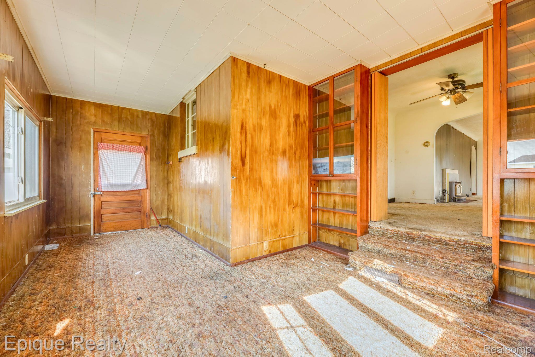 Empty wood-paneled room with broad windows, built-in shelves, and a short step into a larger room.