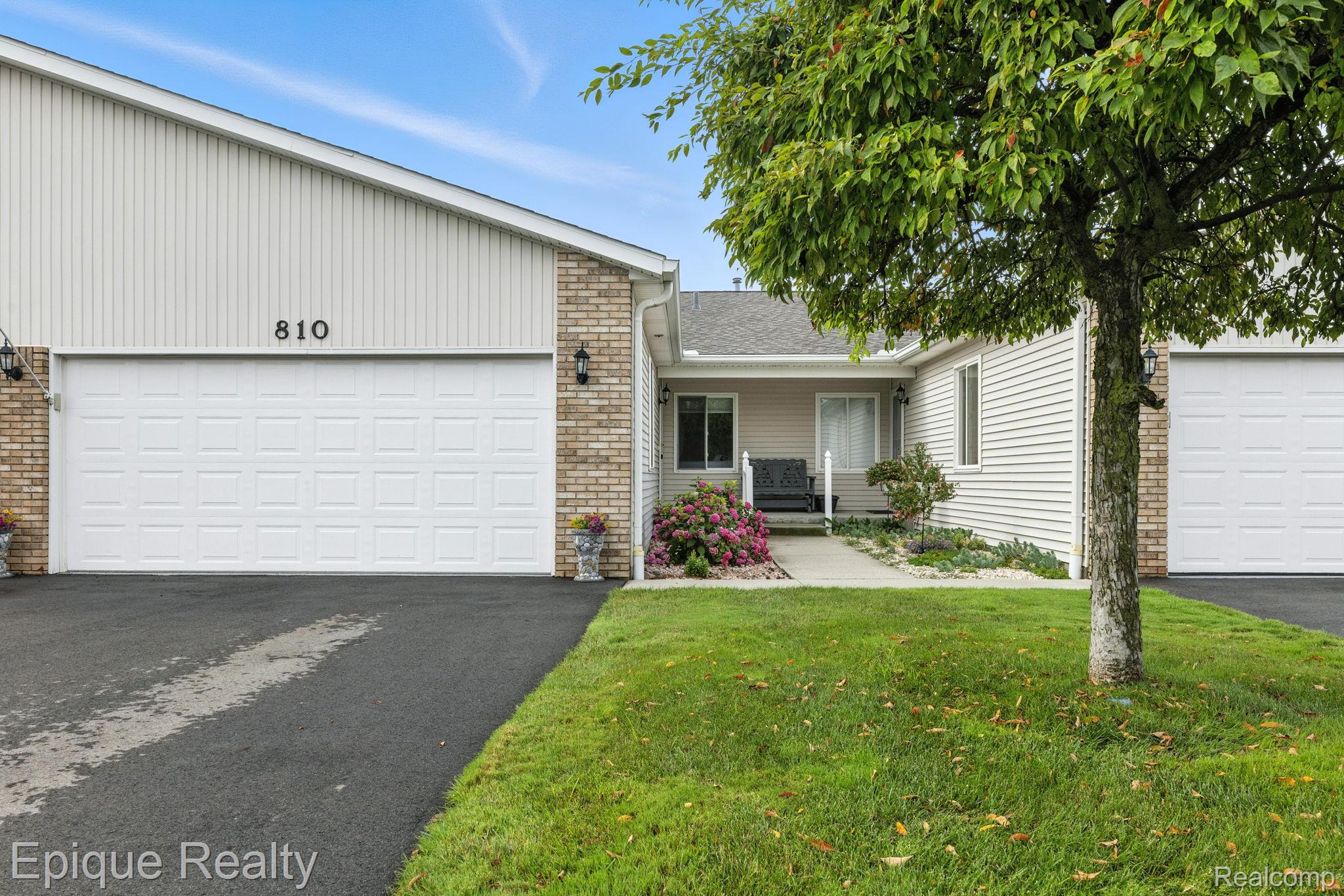 Home exterior with white garage doors, brick trim, and a landscaped entry beneath a leafy tree.