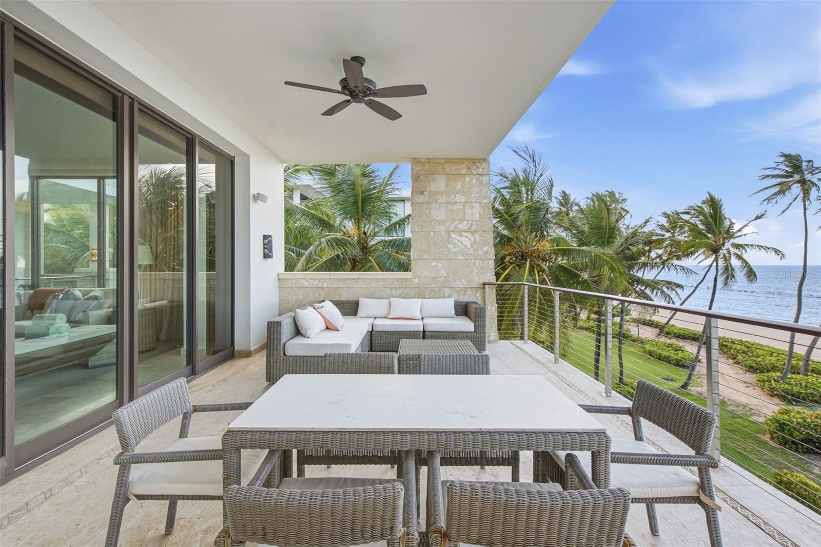 Covered terrace with wicker dining and lounge furniture overlooking palm trees, a sandy beach, and the ocean.