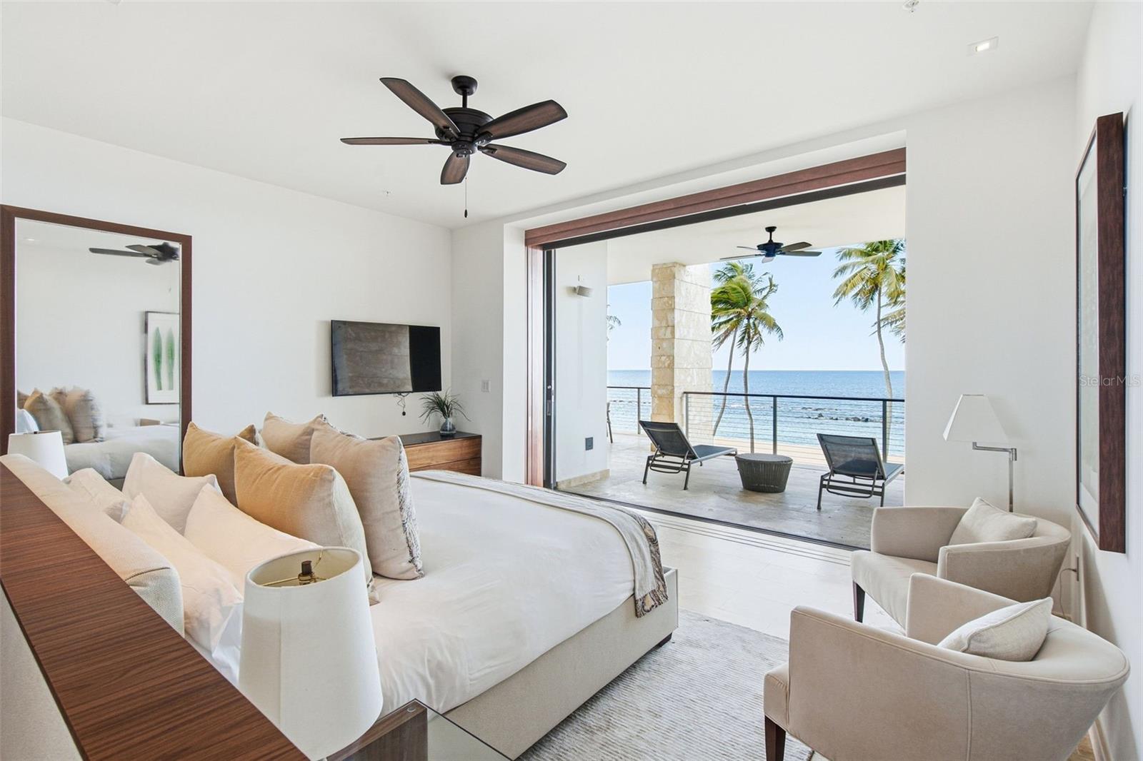 Furnished bedroom with a king bed and open balcony overlooking the ocean and palm trees.