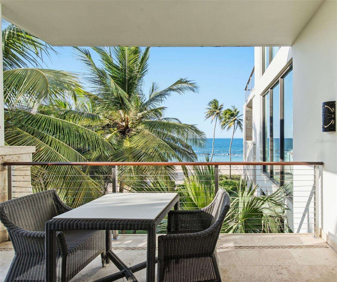 Furnished balcony overlooking palm trees and ocean, with wicker dining set.