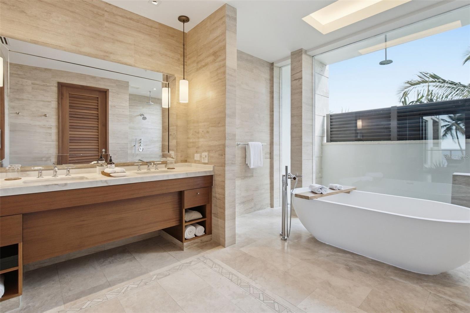 Modern stone-tiled bathroom with a long double vanity and freestanding tub beside large windows.