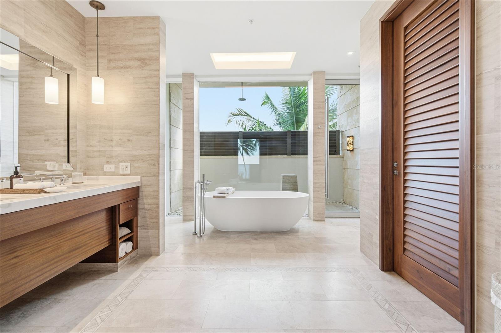Luxury bathroom with a freestanding soaking tub, stone tile, and a large window overlooking palm trees.