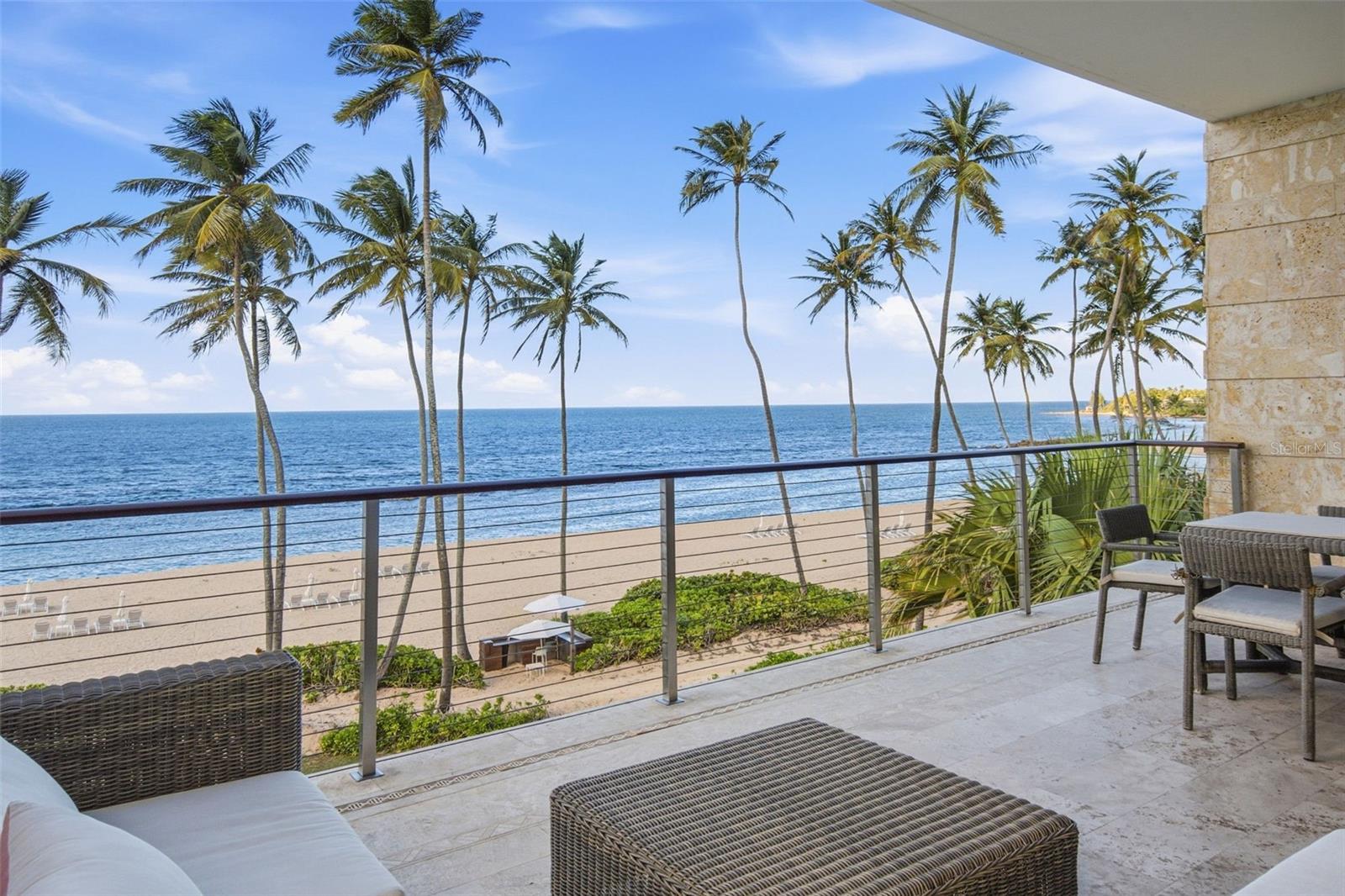 Furnished balcony overlooking a palm-lined beach and ocean, with wicker seating and a glass railing.