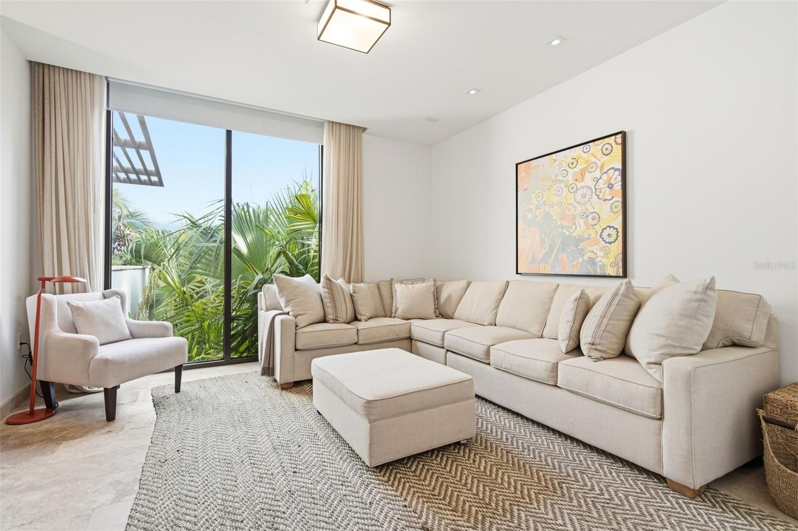 Furnished living room with a cream sectional and armchair beside glass doors overlooking tropical palms.