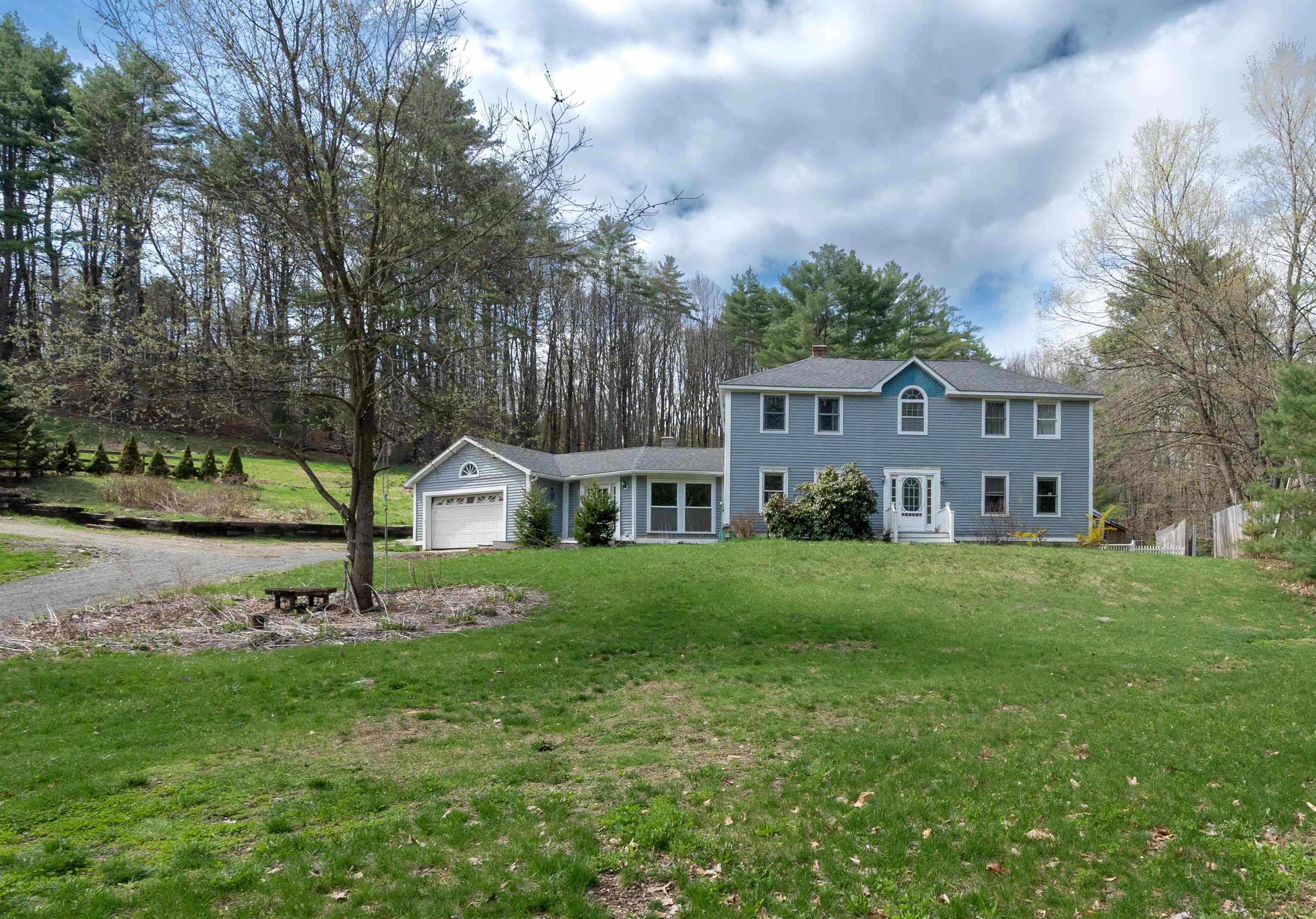 Blue two-story house with an attached garage, broad lawn, and wooded surroundings.