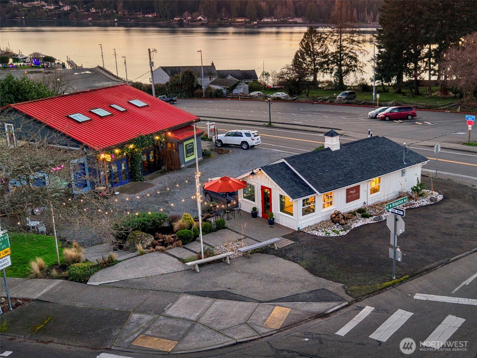 Small white building with a red door and patio umbrella beside a waterfront road.