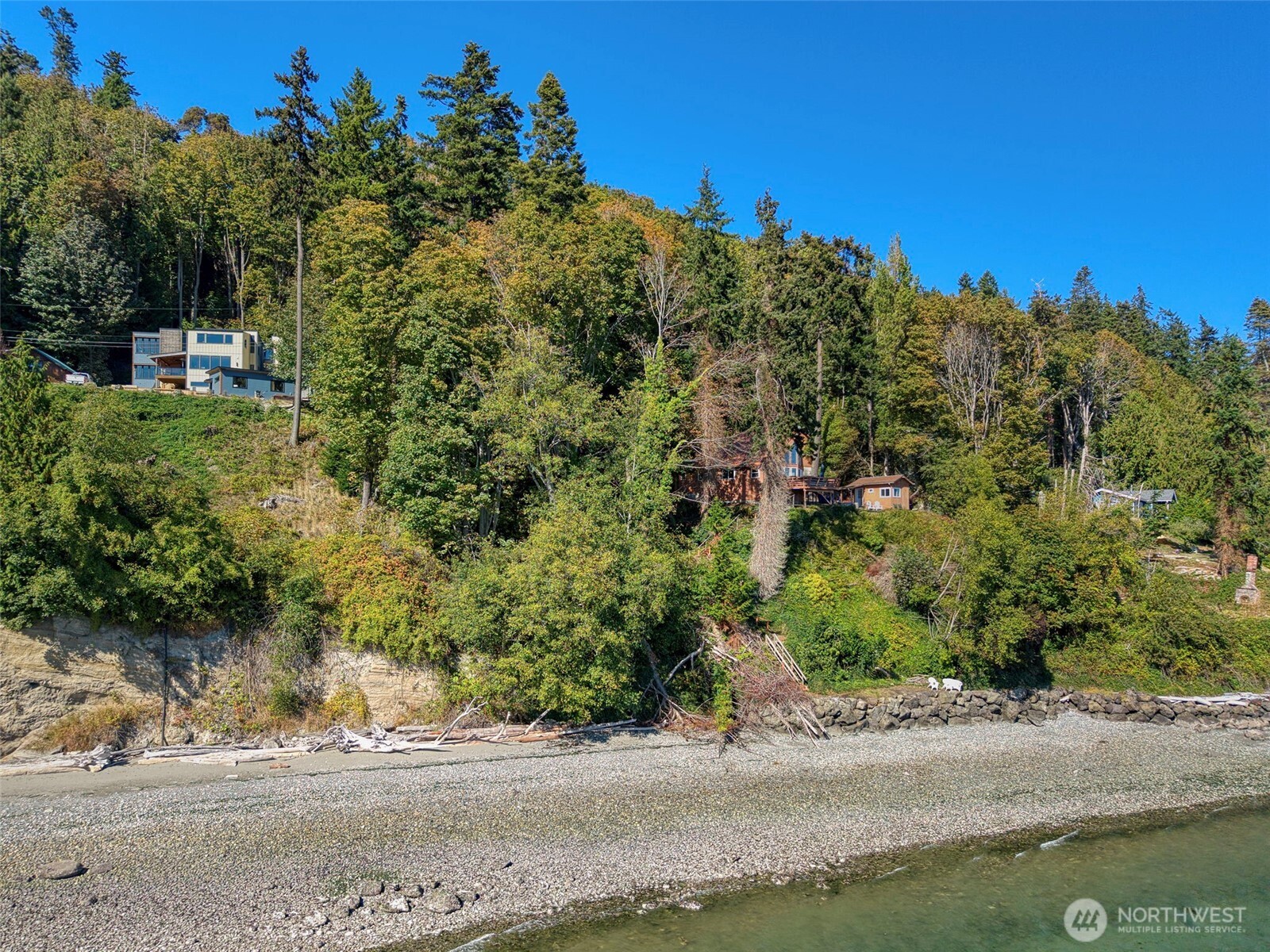 Wooded hillside homes above a pebbled waterfront shoreline.