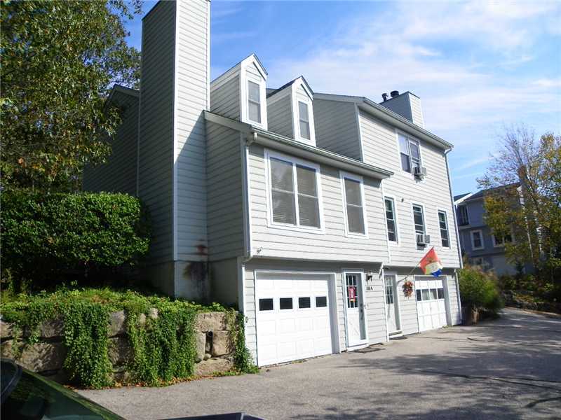 Gray-sided multi-level building with two garage doors, a paved driveway, and a stone retaining wall.